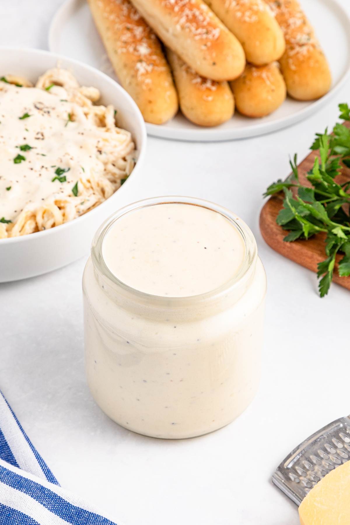 Jar of creamy white sauce on a table with pasta, breadsticks, parsley, and a striped napkin nearby.