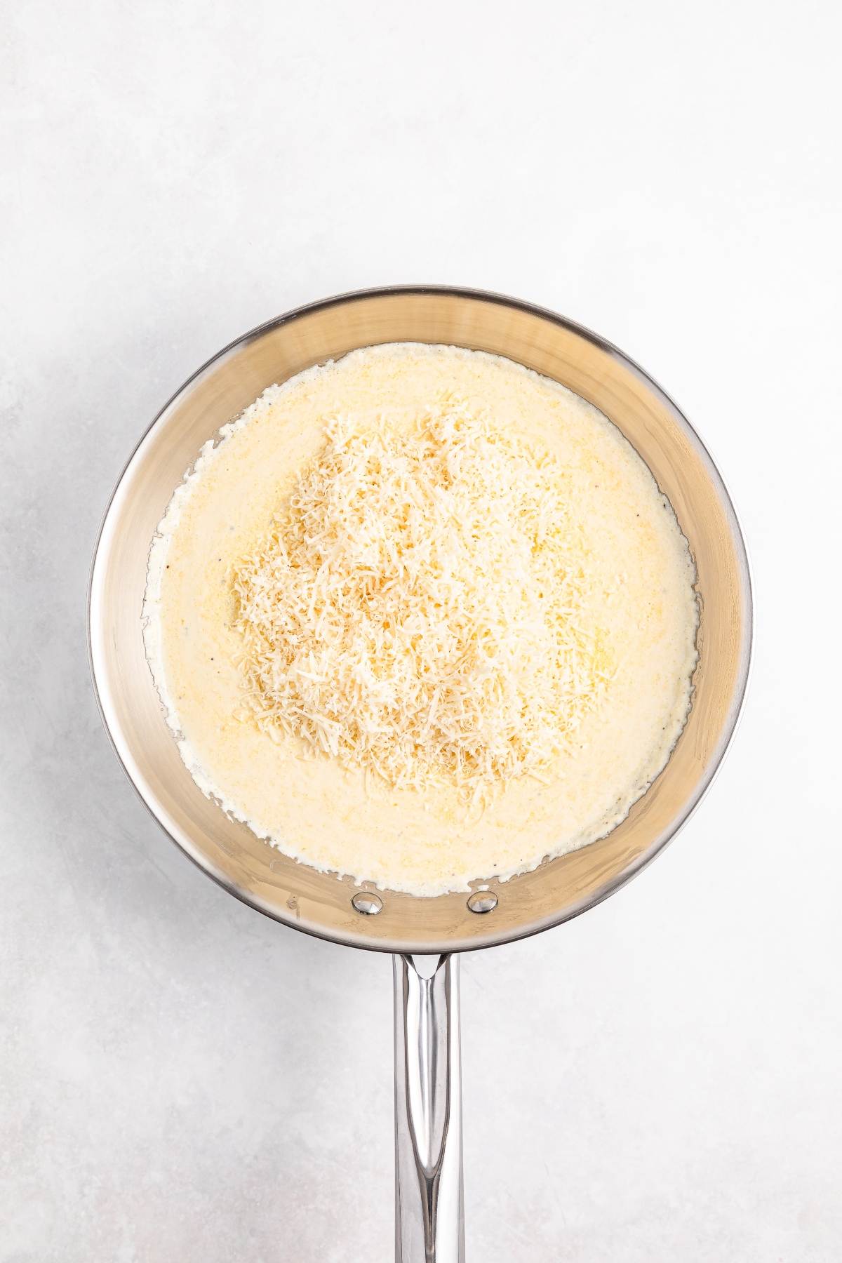 A stainless steel pan with grated cheese atop a creamy white sauce on a light surface.