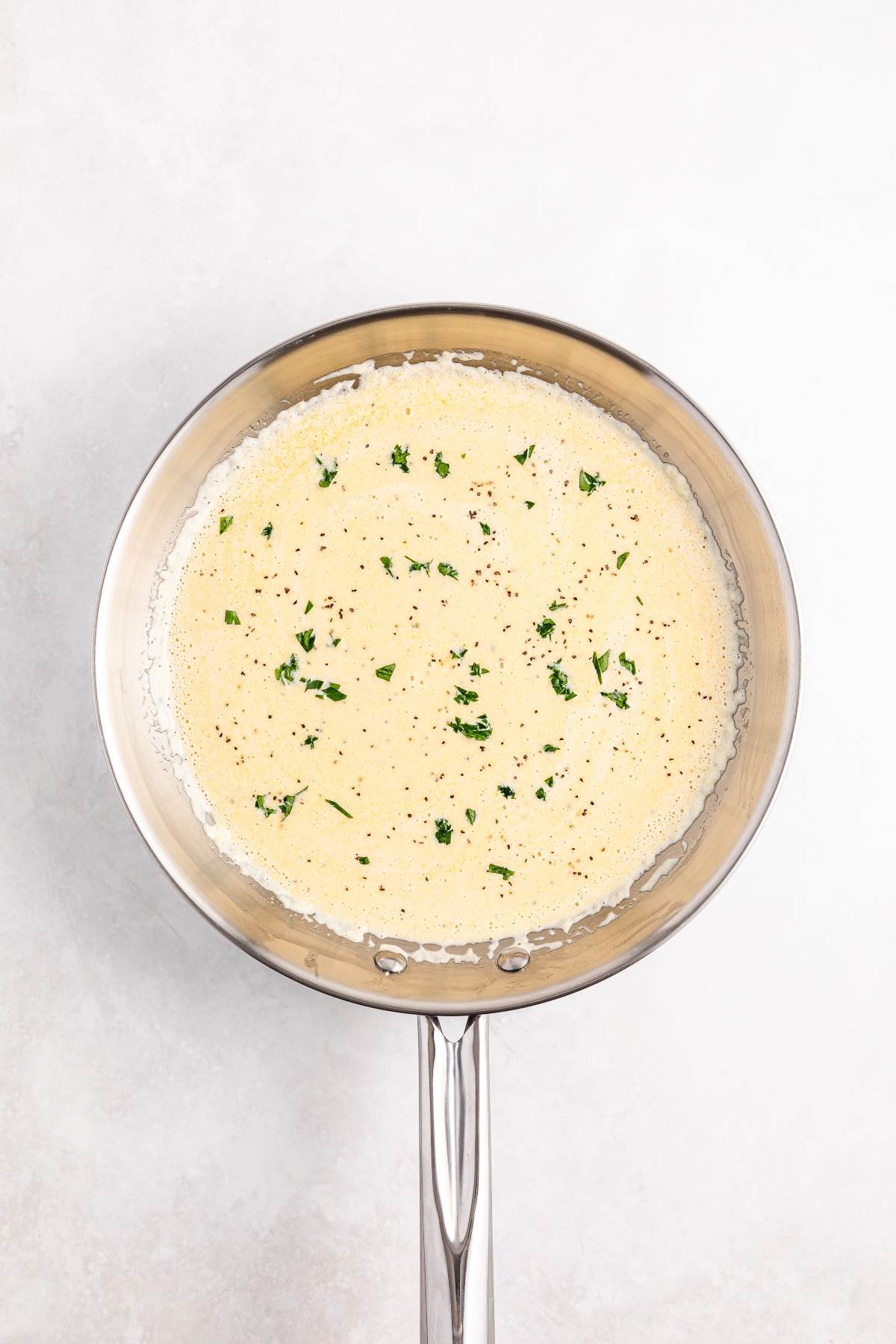 A skillet filled with a creamy, speckled sauce topped with chopped herbs, on a light background.