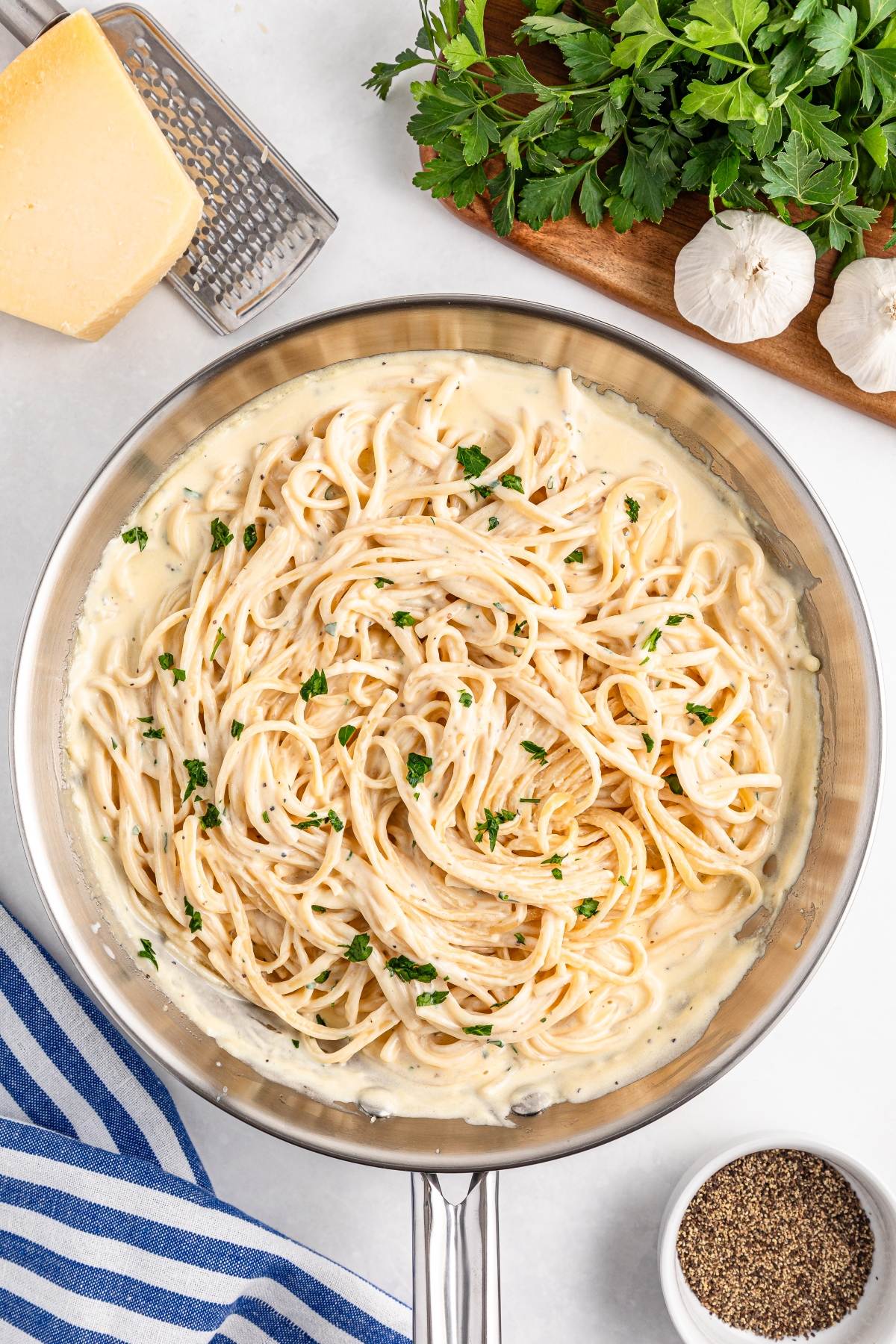Spaghetti in creamy sauce in a pan, garnished with parsley, with cheese, garlic, and herbs nearby.
