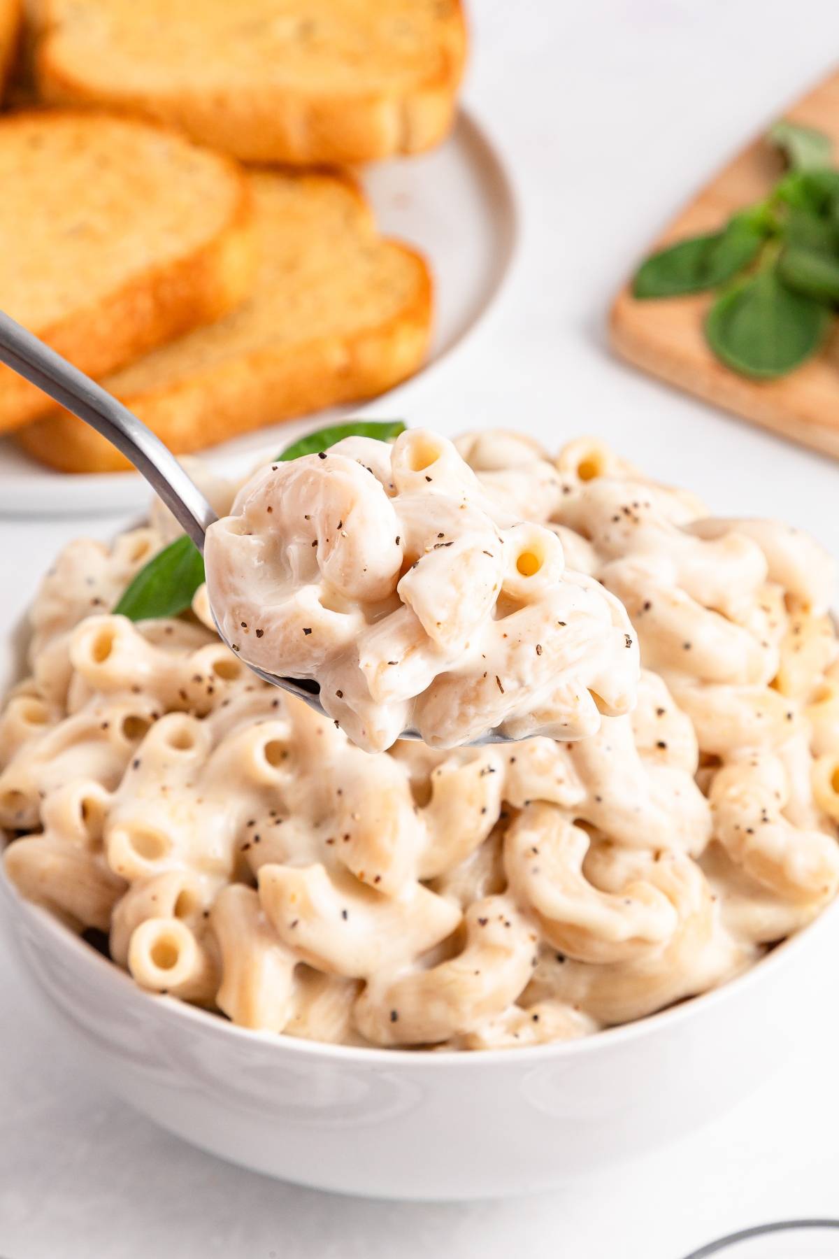 A spoonful of creamy macaroni and cheese above a bowl, with toasted bread in the background.