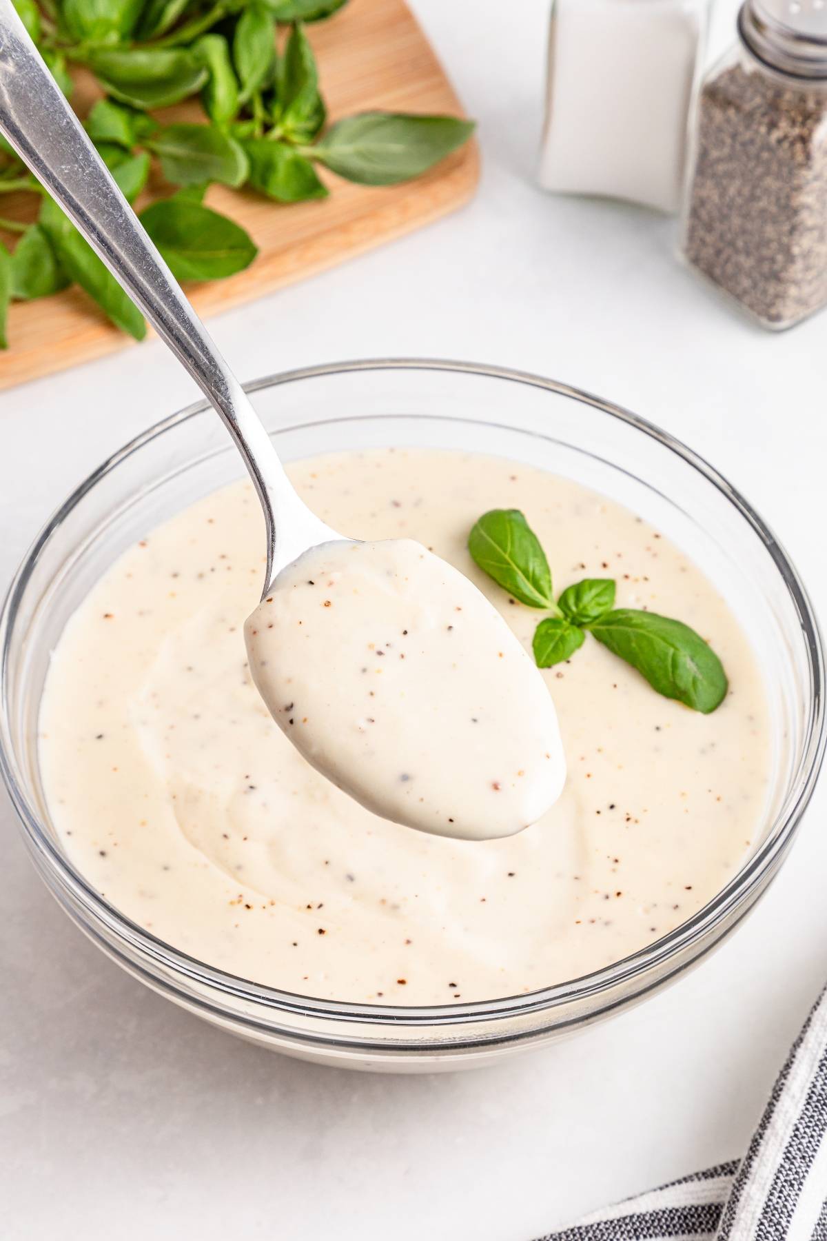 A spoon holding creamy salad dressing above a bowl, garnished with fresh basil leaves.