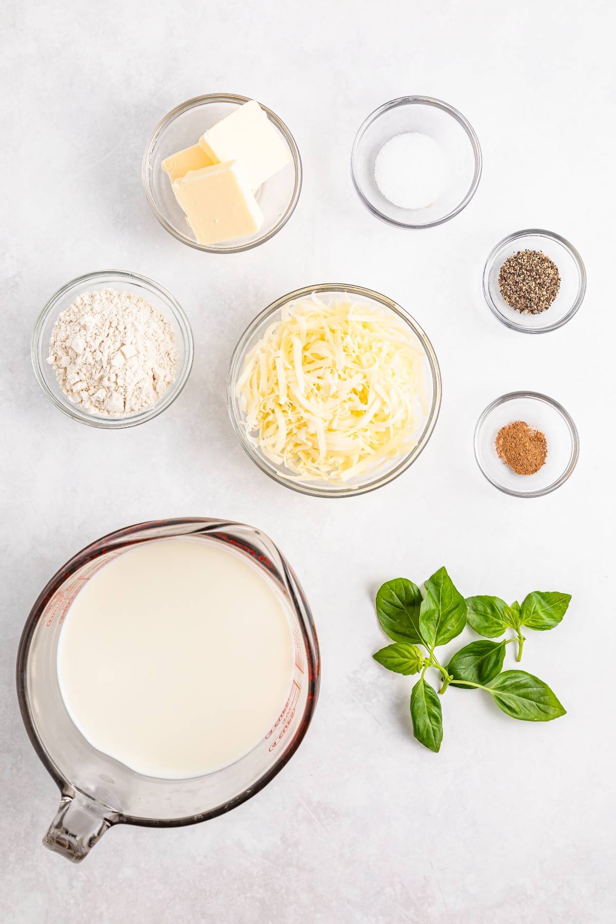 Cheese sauce ingredients: milk, shredded cheese, butter, flour, basil, salt, pepper, and nutmeg on a white surface.