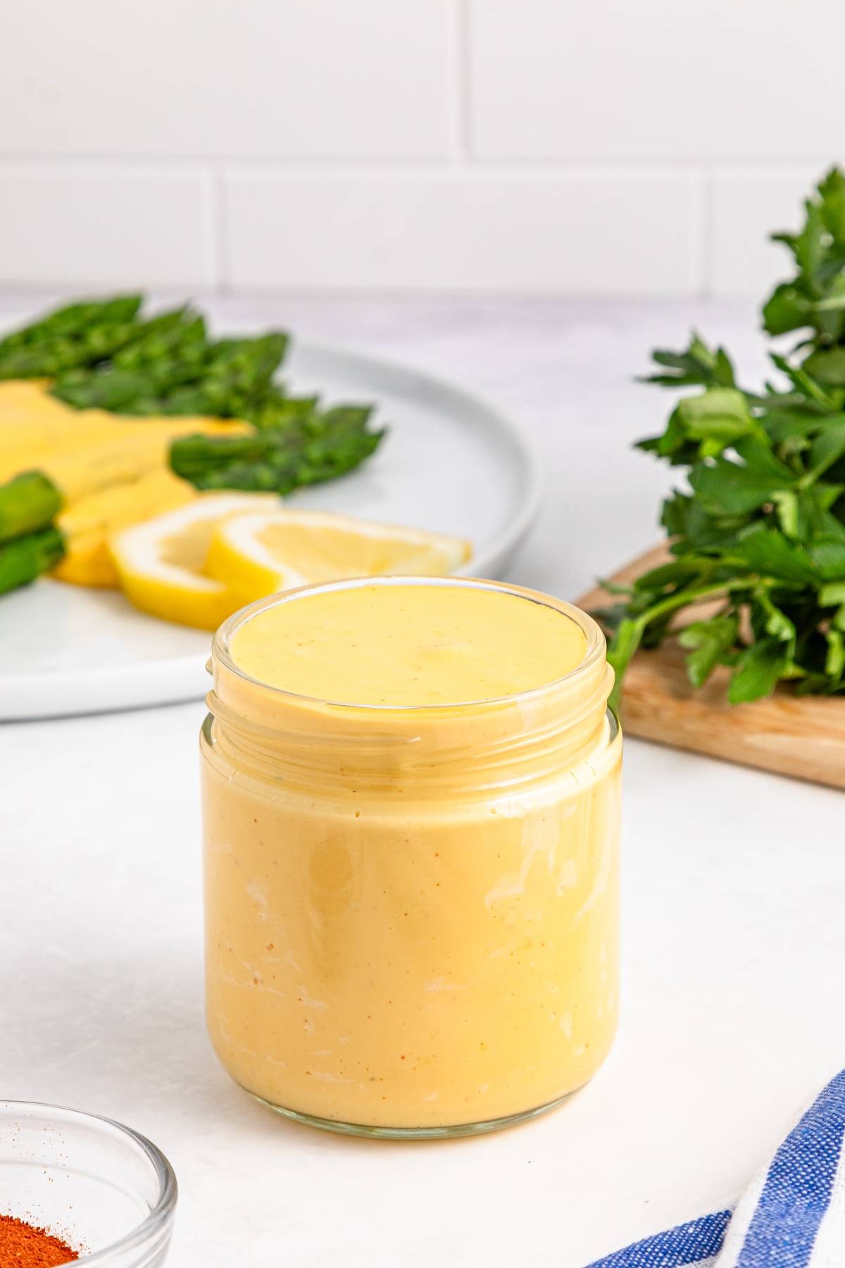 A glass jar filled with creamy yellow sauce, with herbs and sliced lemons in the background.