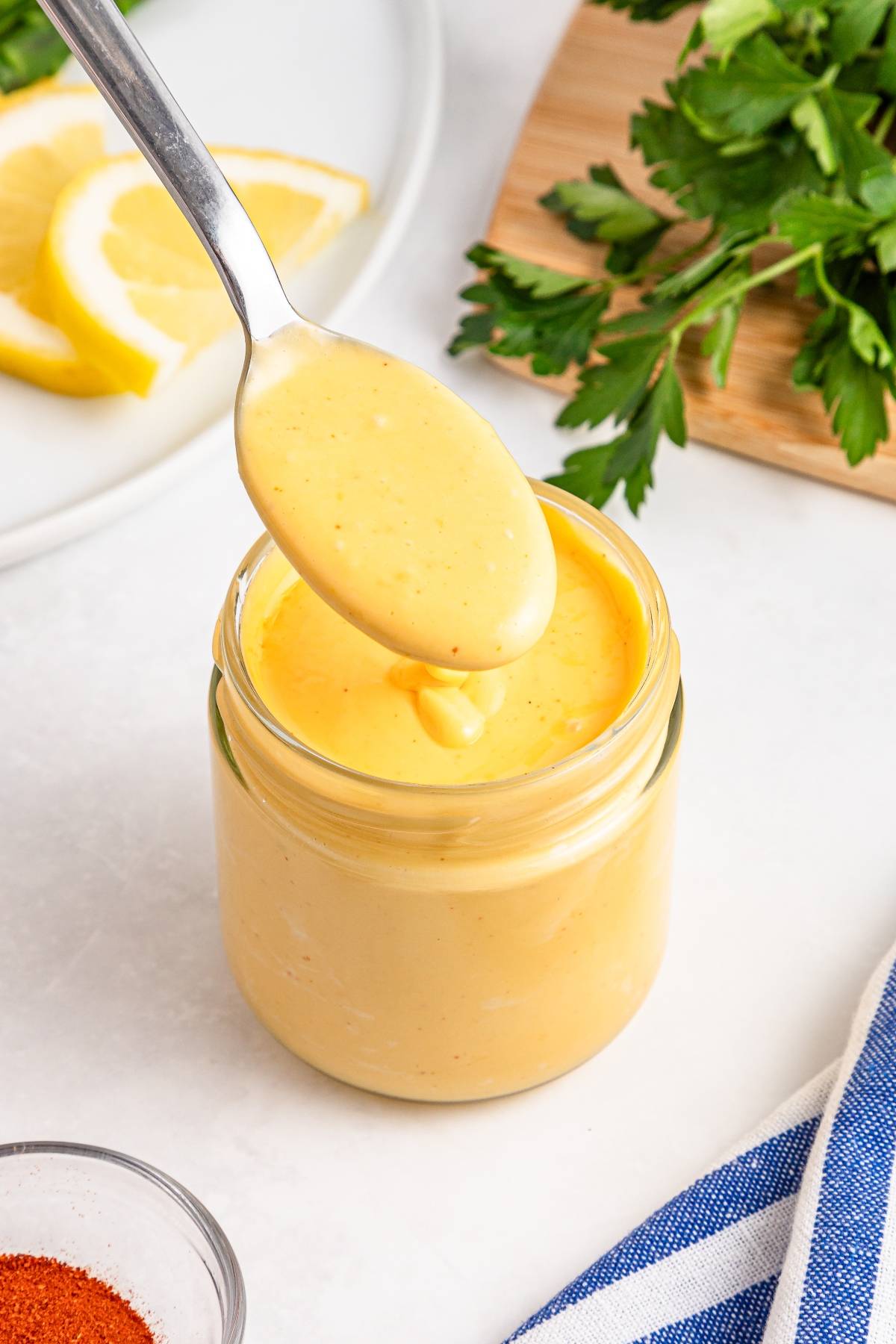 A spoon holding yellow sauce over a glass jar, with lemon slices and parsley in the background.
