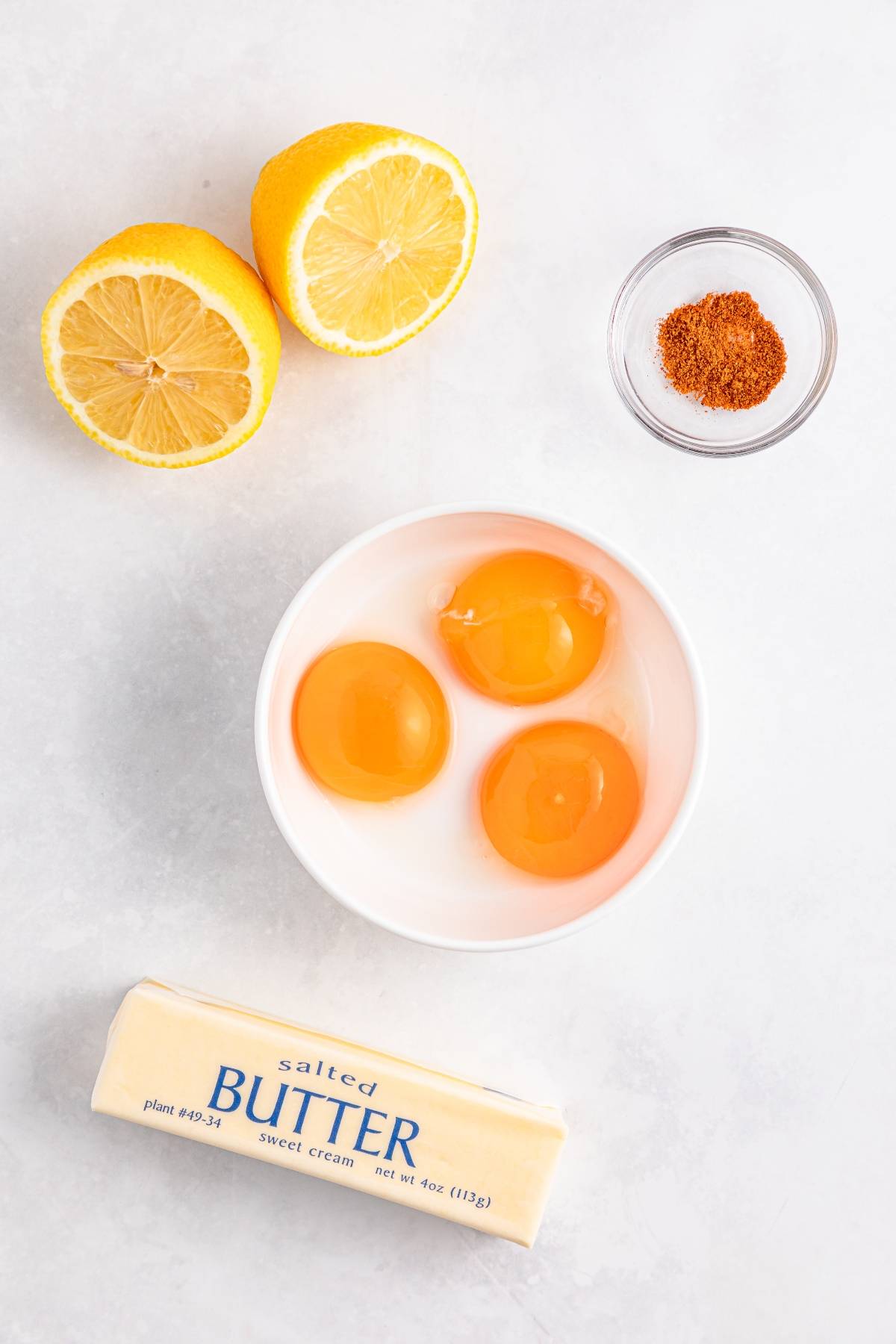 Three egg yolks in a bowl, a stick of butter, two lemon halves, and a small bowl of ground spice.