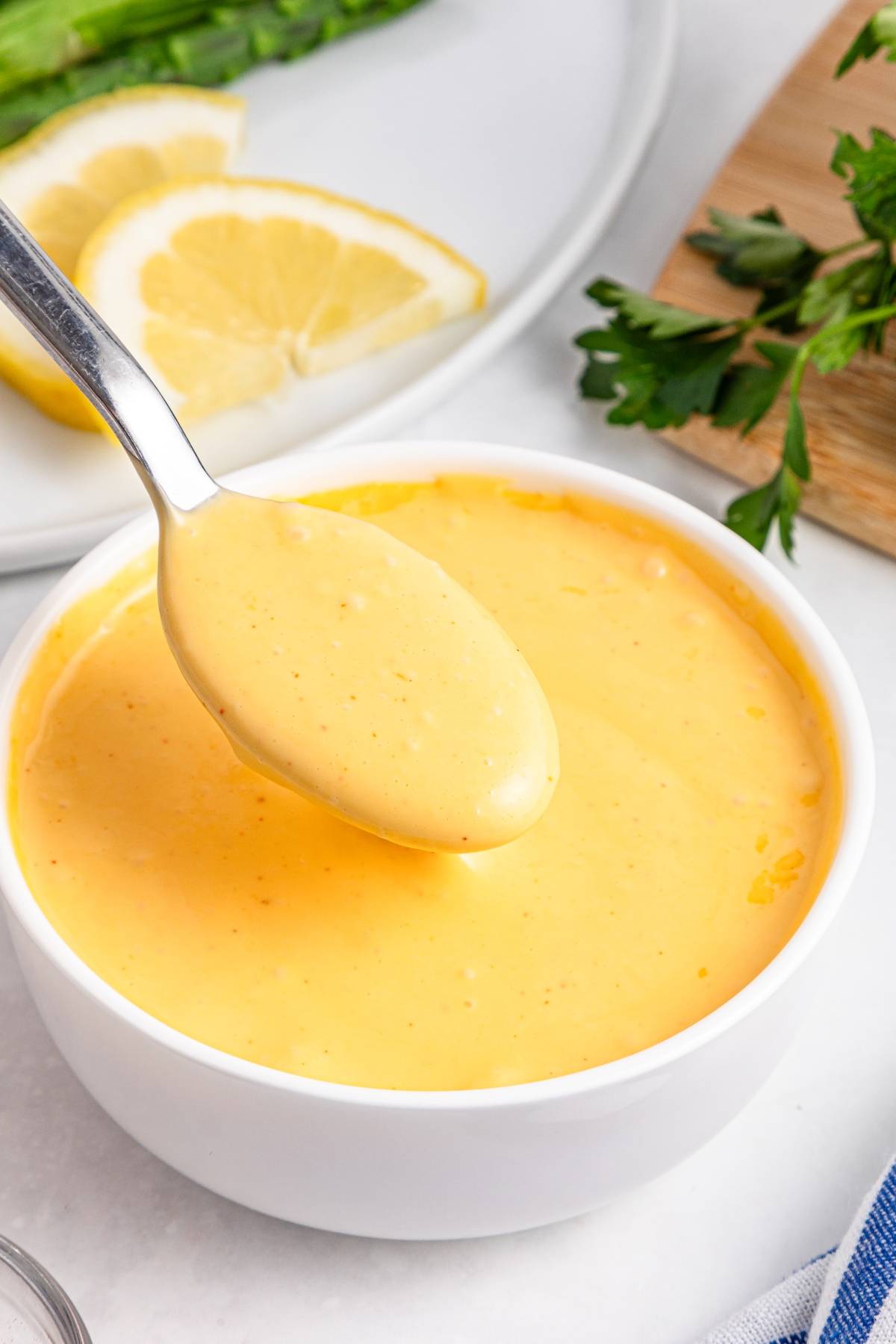 A spoon holding creamy yellow sauce above a white bowl, with lemon slices and herbs in the background.