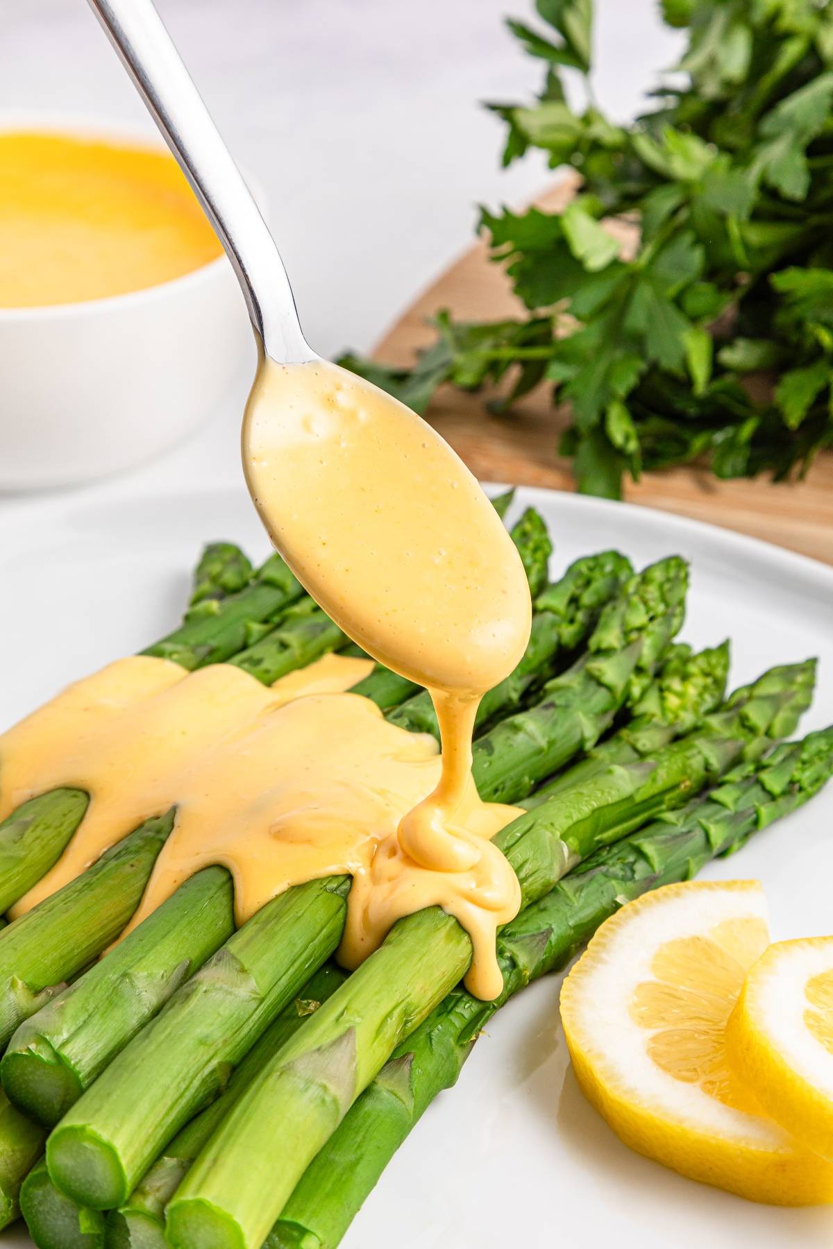 A spoon drizzling hollandaise sauce over steamed asparagus, with lemon slices and parsley on the side.