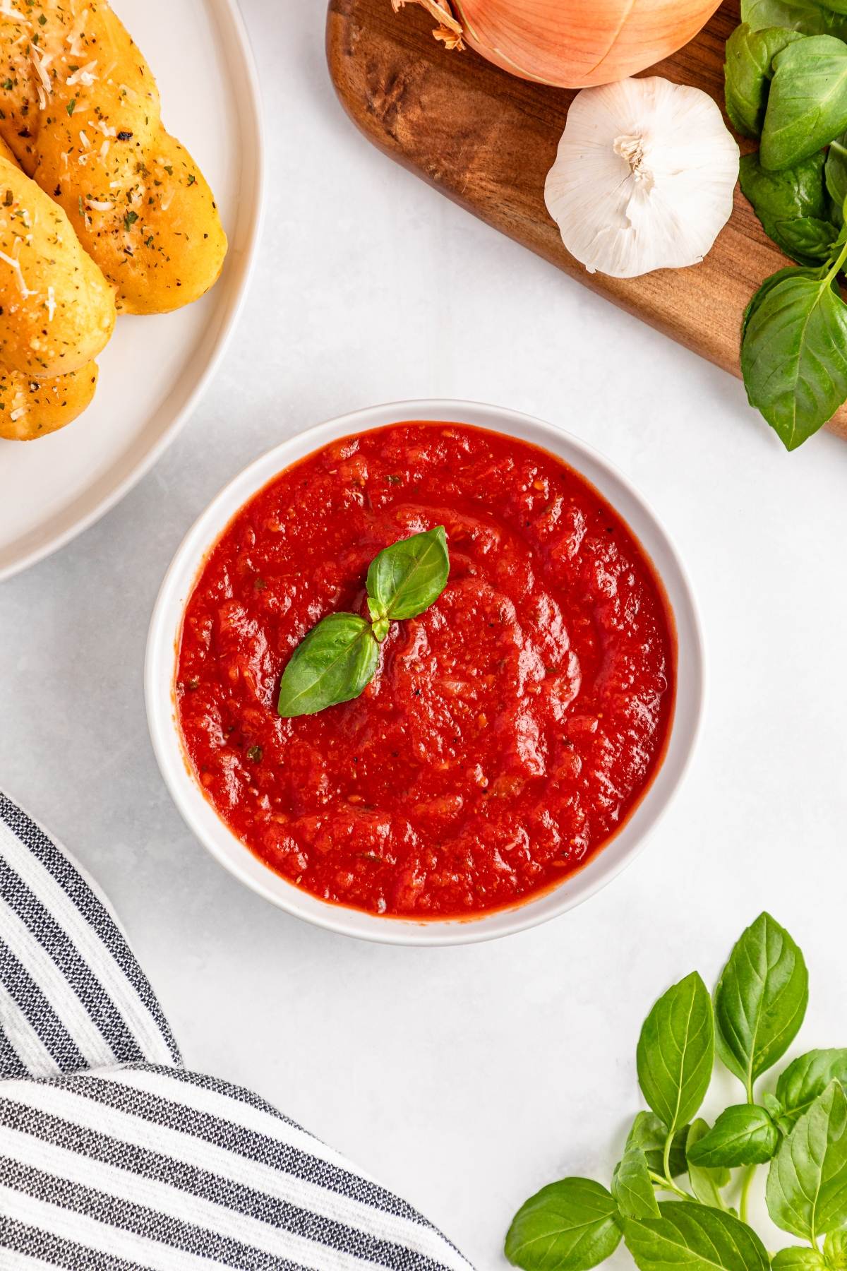 Bowl of red marinara sauce garnished with basil, surrounded by garlic, onion, basil leaves, and breadsticks.