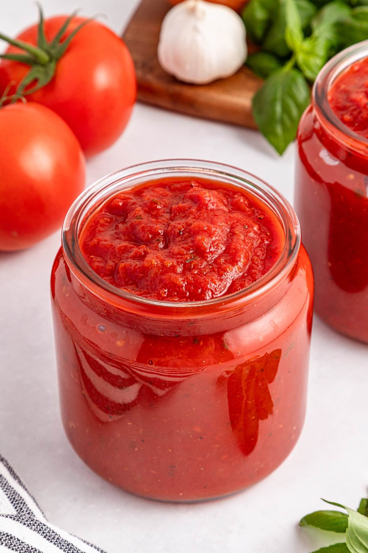 A jar of tomato sauce with fresh tomatoes, garlic, and basil in the background on a white surface.