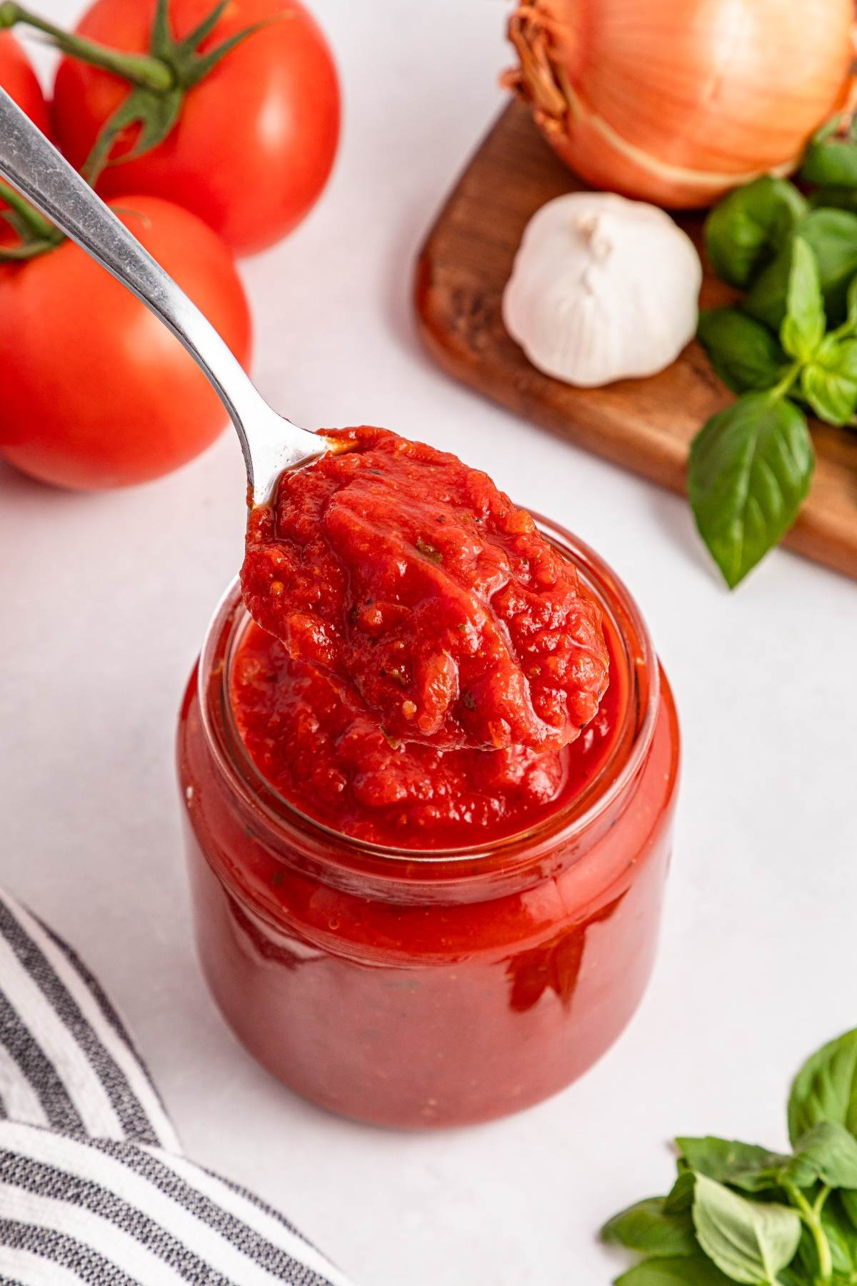 Spoonful of tomato sauce held above a jar, with tomatoes, garlic, onion, and basil in the background.