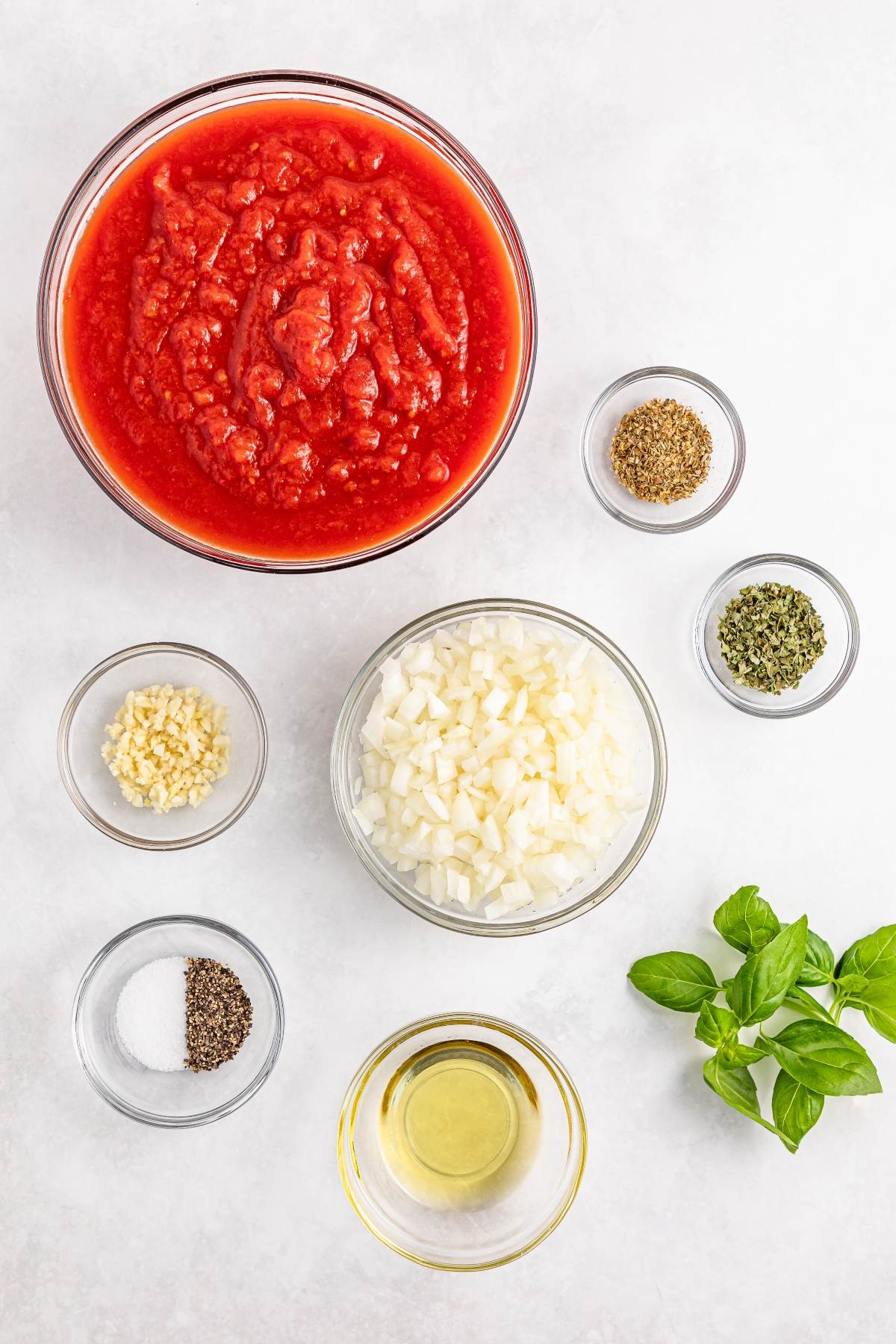 Bowls of crushed tomatoes, chopped onions, minced garlic, oil, herbs, salt, pepper, and fresh basil on a white surface.