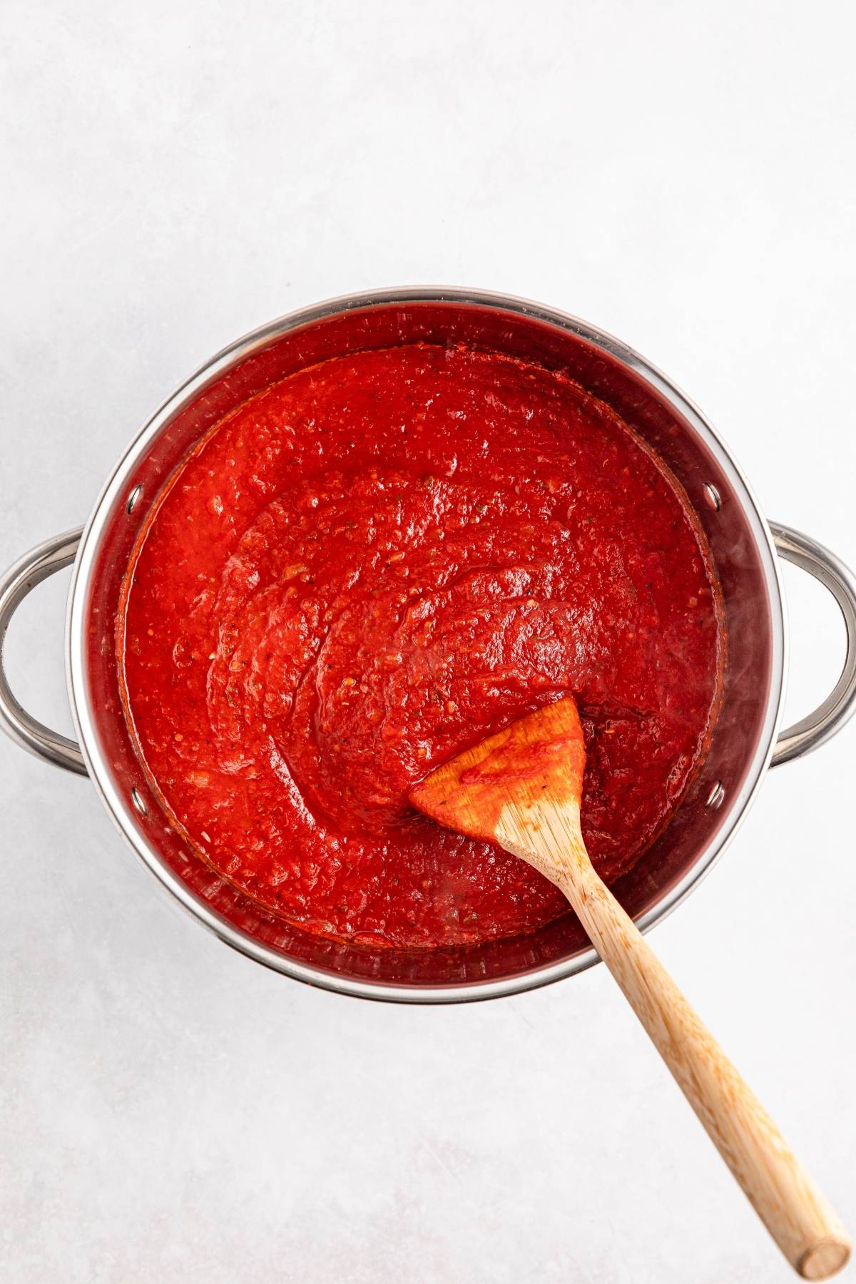 A pot of red tomato sauce being stirred with a wooden spoon on a white background.