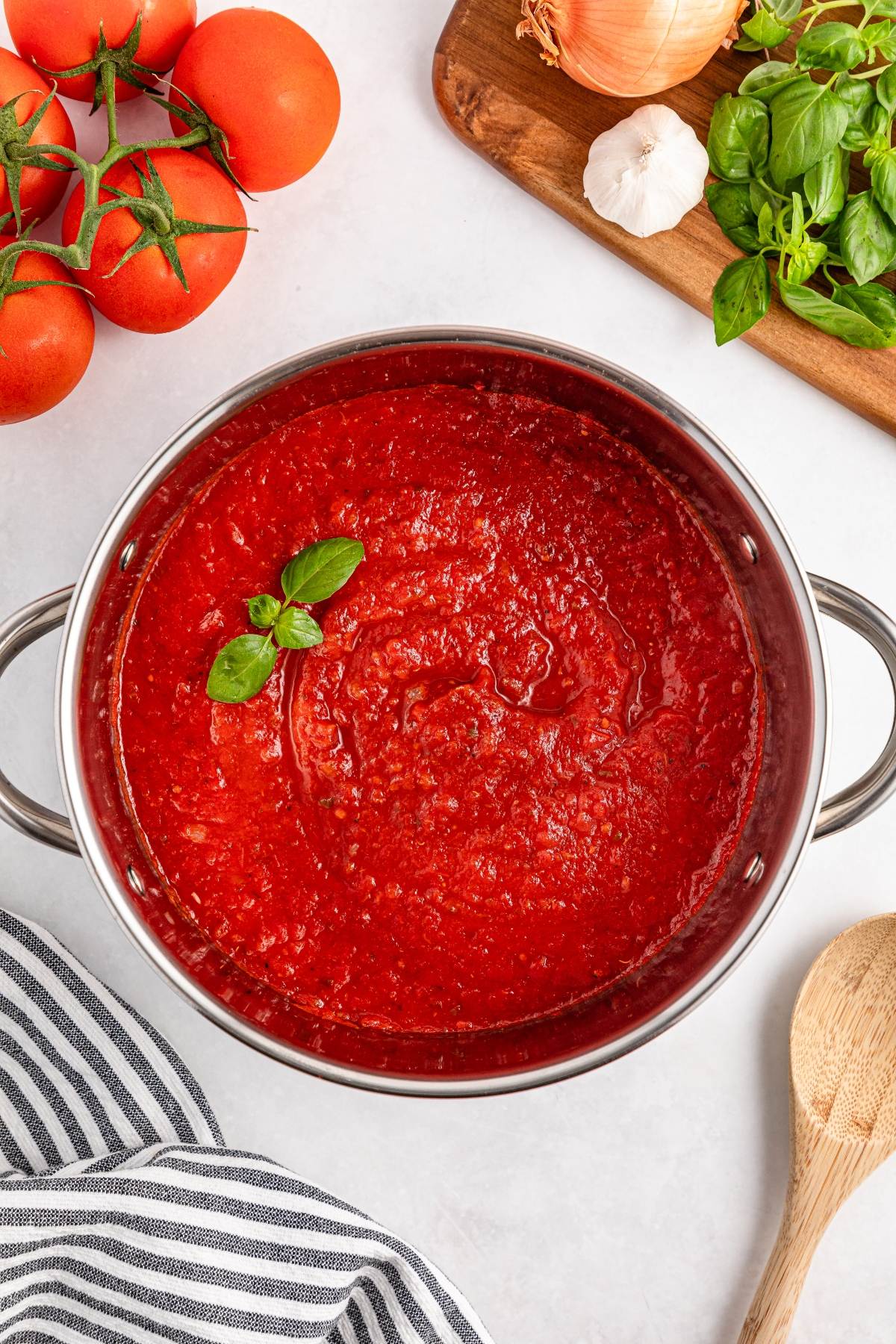 A pot of tomato sauce garnished with basil, surrounded by fresh tomatoes, onion, garlic, and basil.