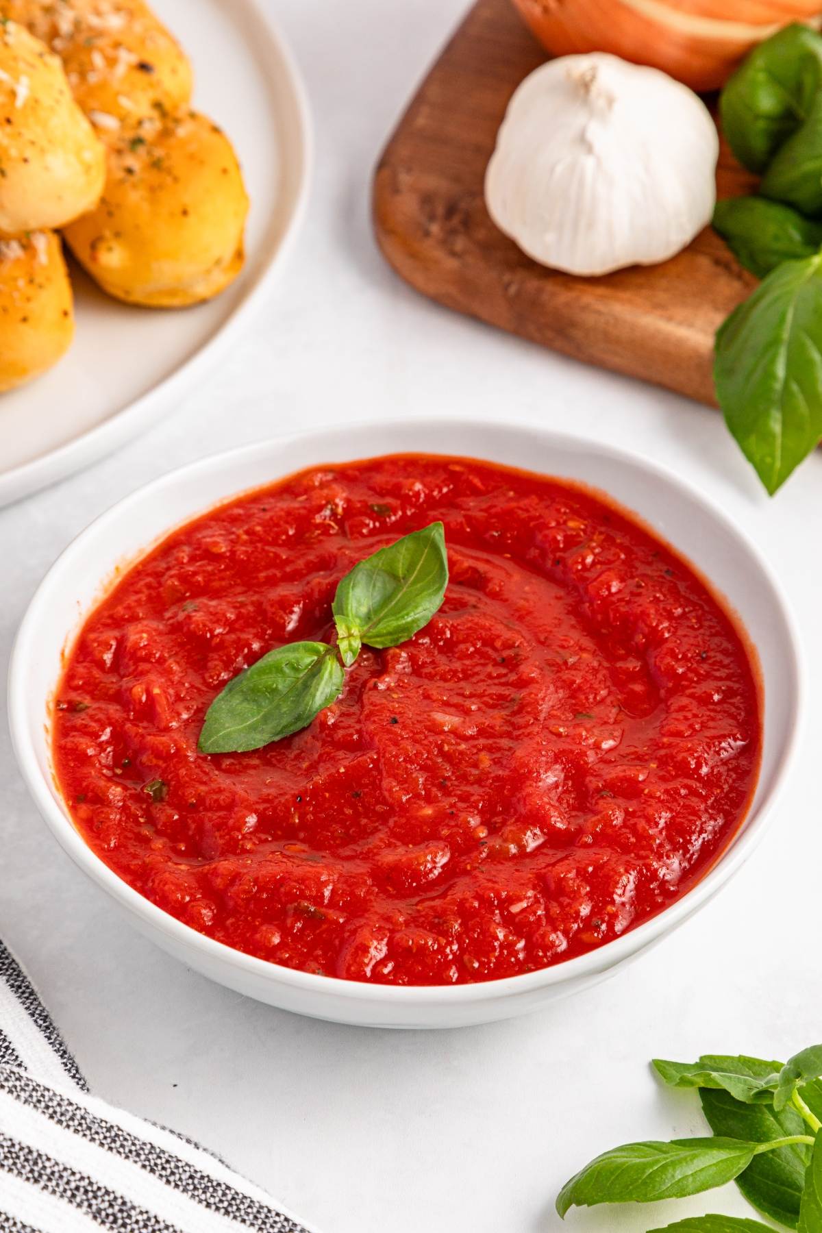 Bowl of tomato sauce garnished with basil, with garlic and breadsticks in the background.