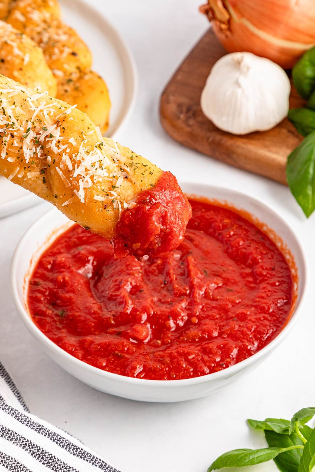 A breadstick dipped in marinara sauce, with garlic, onion, and basil in the background.