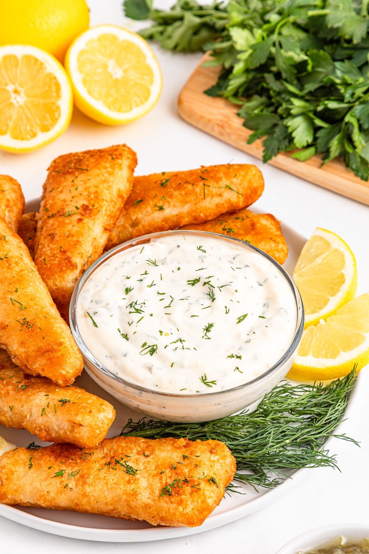Fried fish sticks with easy tartar sauce, lemon slices, and fresh herbs on a white plate.