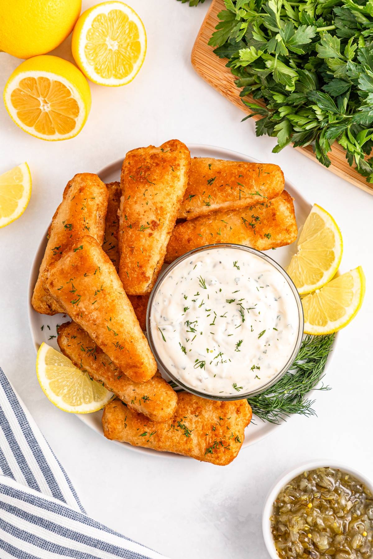 Plate of breaded fish fillets with lemon wedges and a bowl of tartar sauce, surrounded by herbs and lemon halves.
