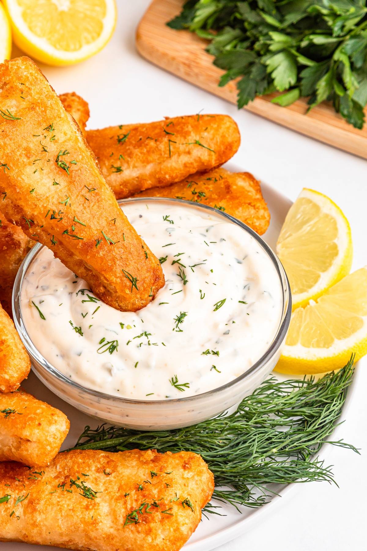 Fried fish sticks with dill dip, lemon wedges, and fresh herbs on a white plate.