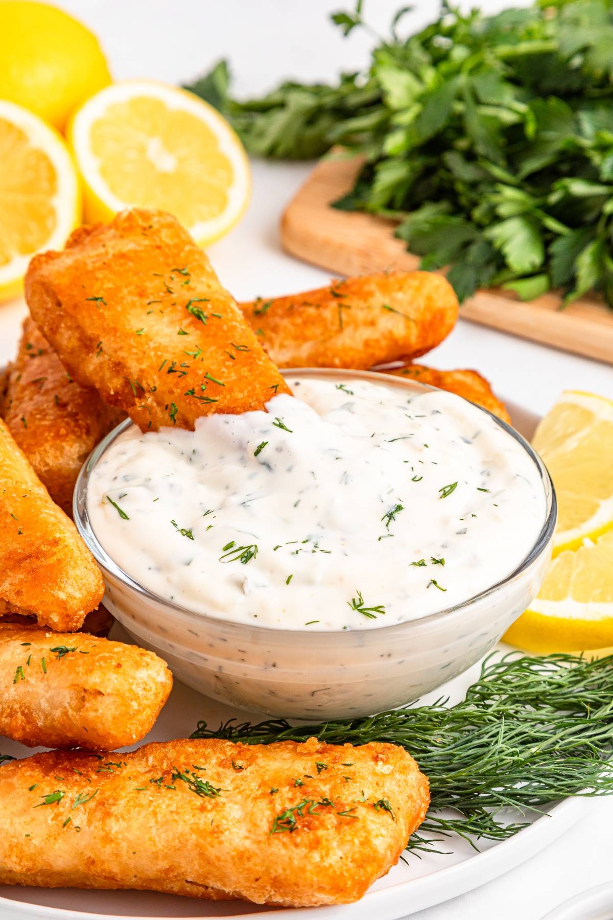 Fried fish sticks with creamy herb dip, fresh dill, lemon slices, and parsley on a plate.