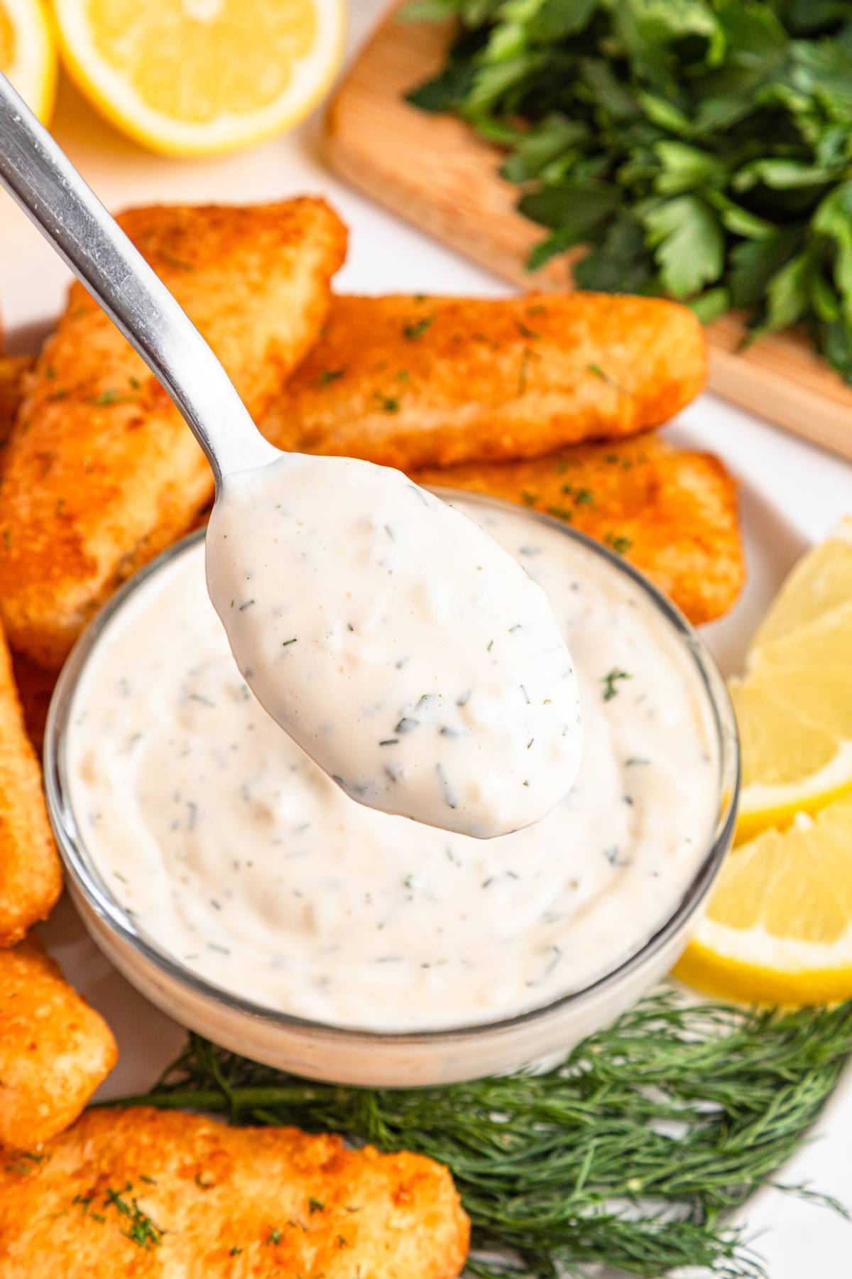 A spoonful of creamy tartar sauce held above a bowl, surrounded by fish sticks, lemon slices, and herbs.
