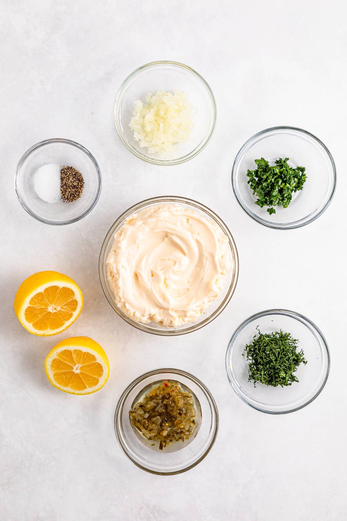 Bowls of mayonnaise, chopped herbs, onions, relish, salt and pepper, and two lemon halves on a white surface.