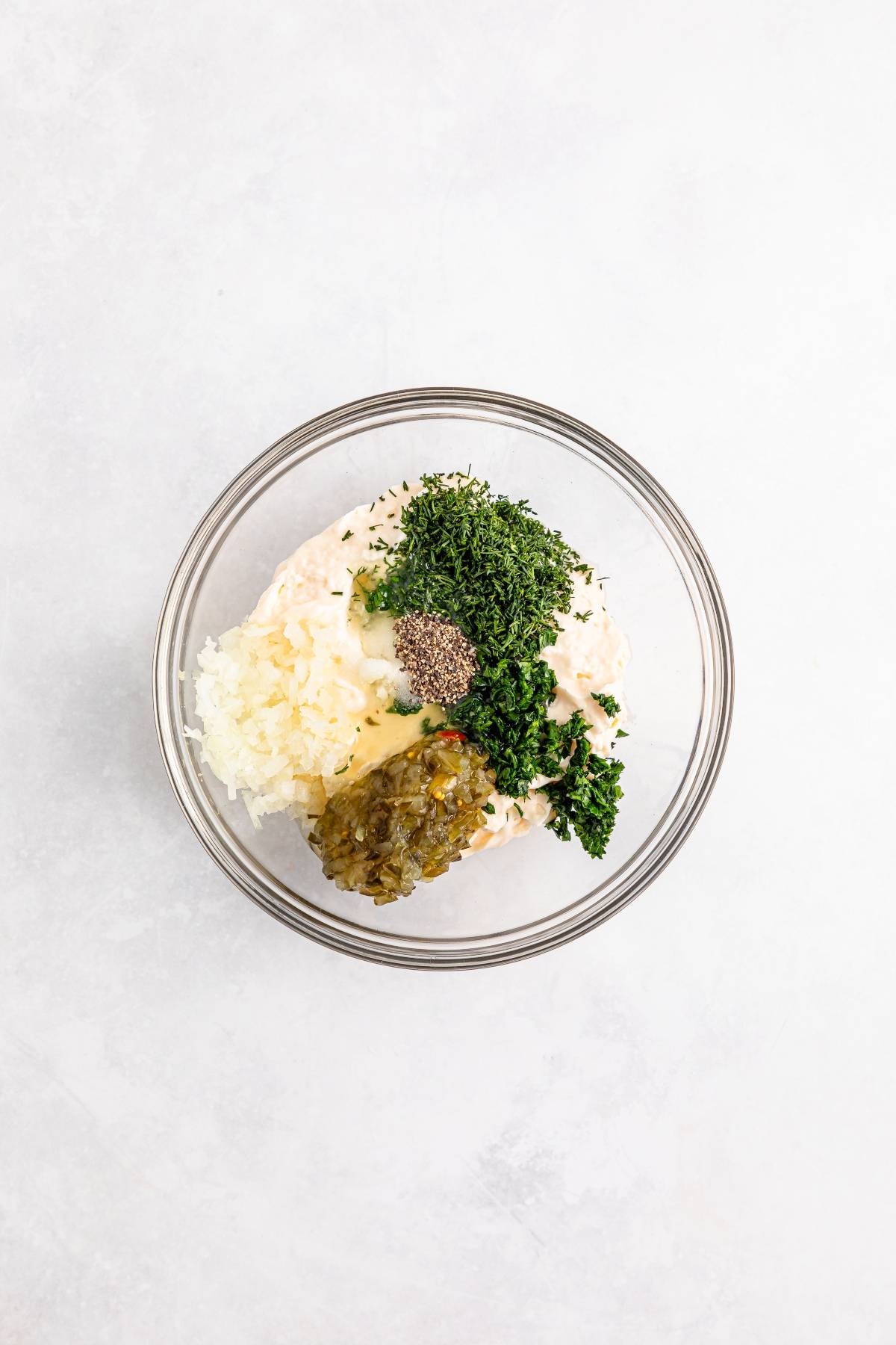 A glass bowl with mayonnaise, chopped herbs, relish, grated onion, and pepper on a white surface.