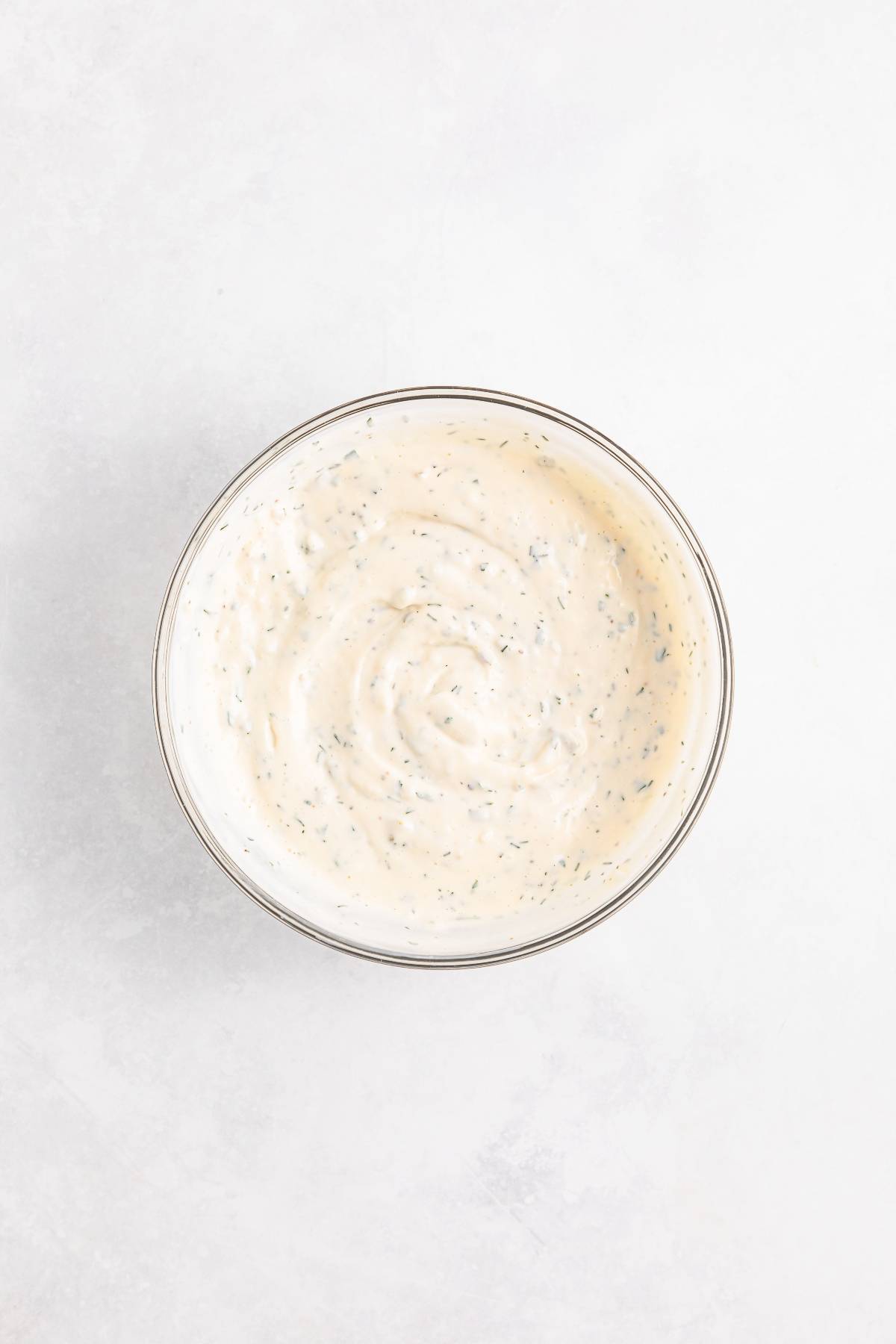 A bowl of creamy white ranch dressing with visible herbs, viewed from above on a light background.