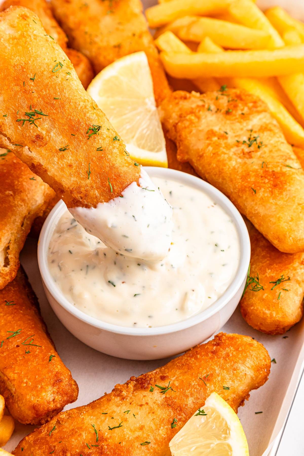 Crispy fried fish is dipped in creamy tartar sauce, surrounded by fries and lemon wedges.