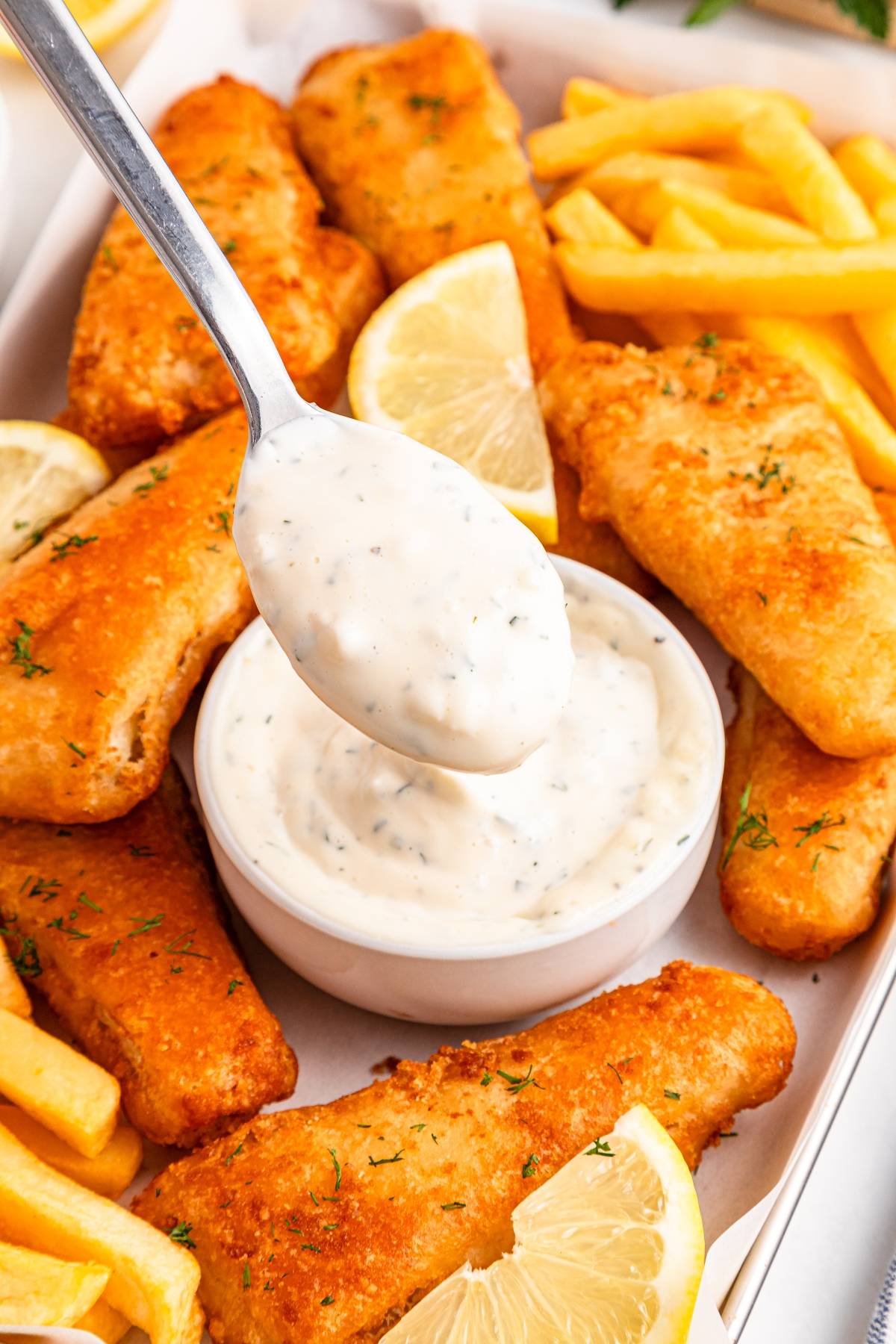 A spoon dips into tartar sauce, surrounded by fried fish, lemon slices, and French fries.