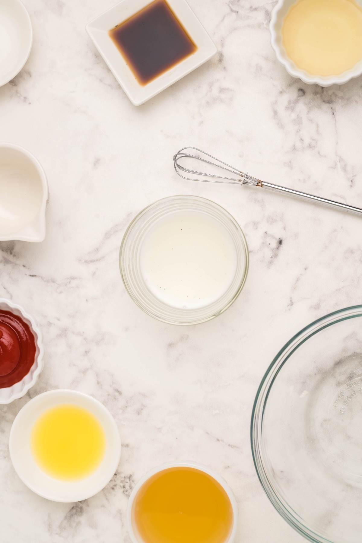 A selection of small bowls filled with homemade sauce recipes, like sweet and sour sauce and egg roll sauce, arranged with a whisk on a white marble surface.