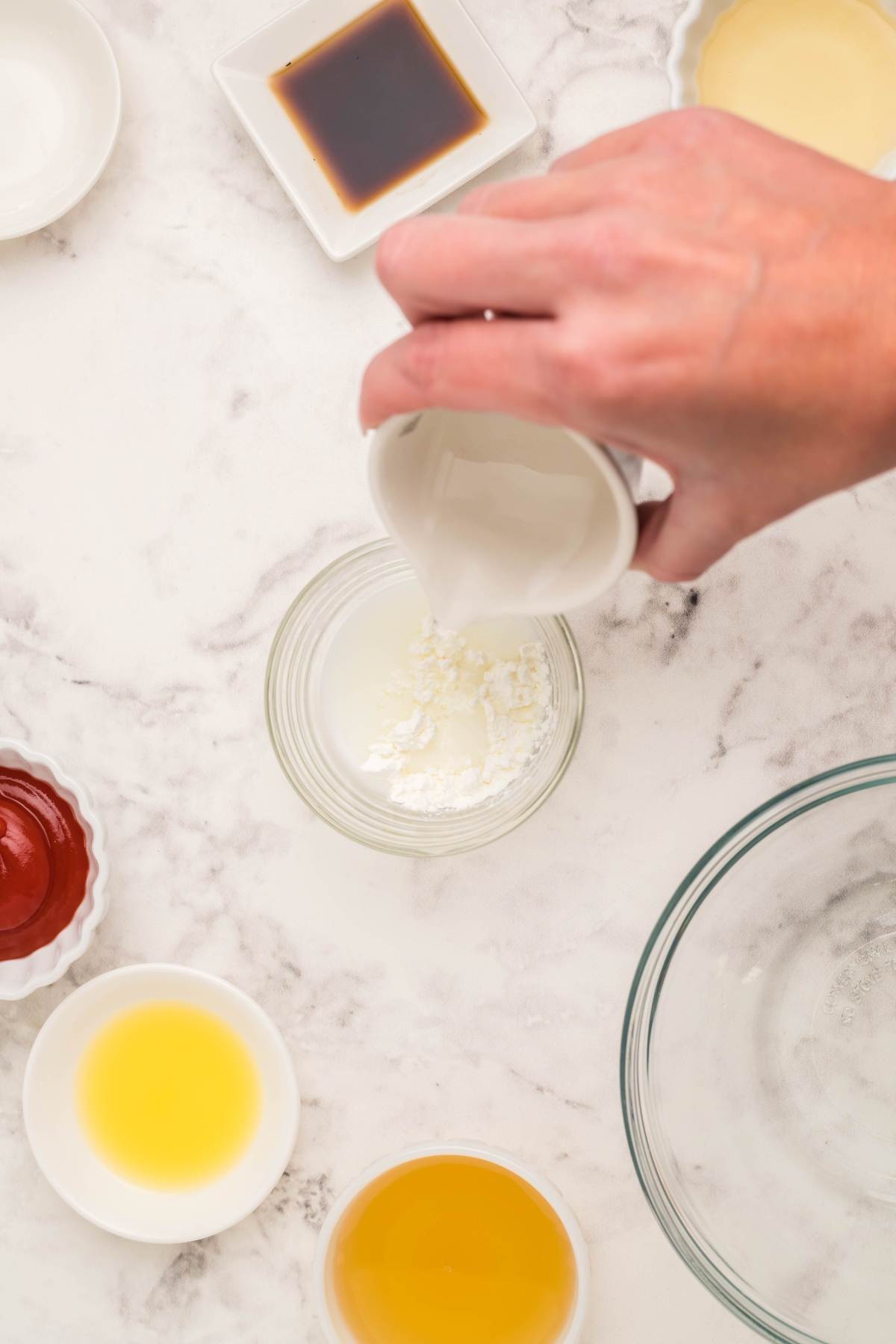 Hand pouring liquid into a small bowl of white powder, surrounded by bowls of sauces like sweet and sour sauce on a marble surface.