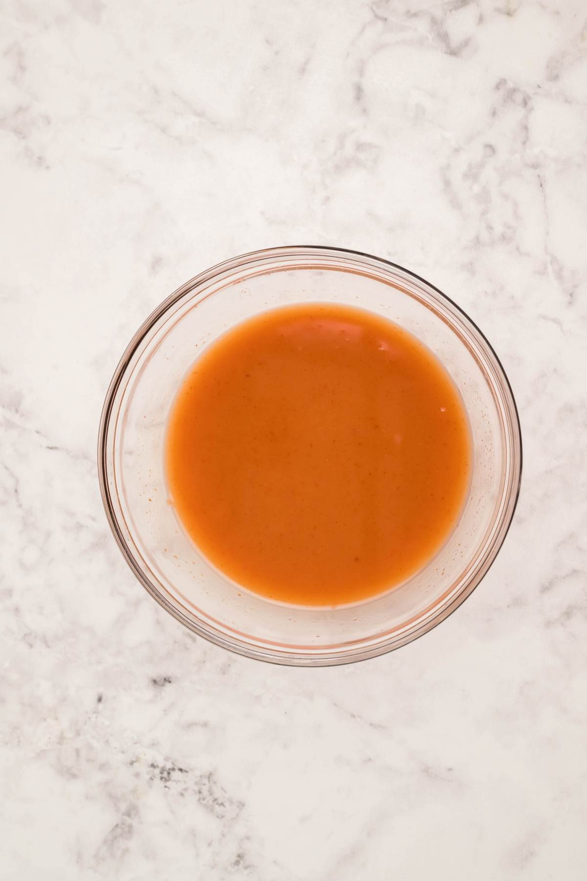 A glass bowl filled with sweet and sour sauce sits on a white marble surface, perfect for those seeking a homemade sauce recipe.