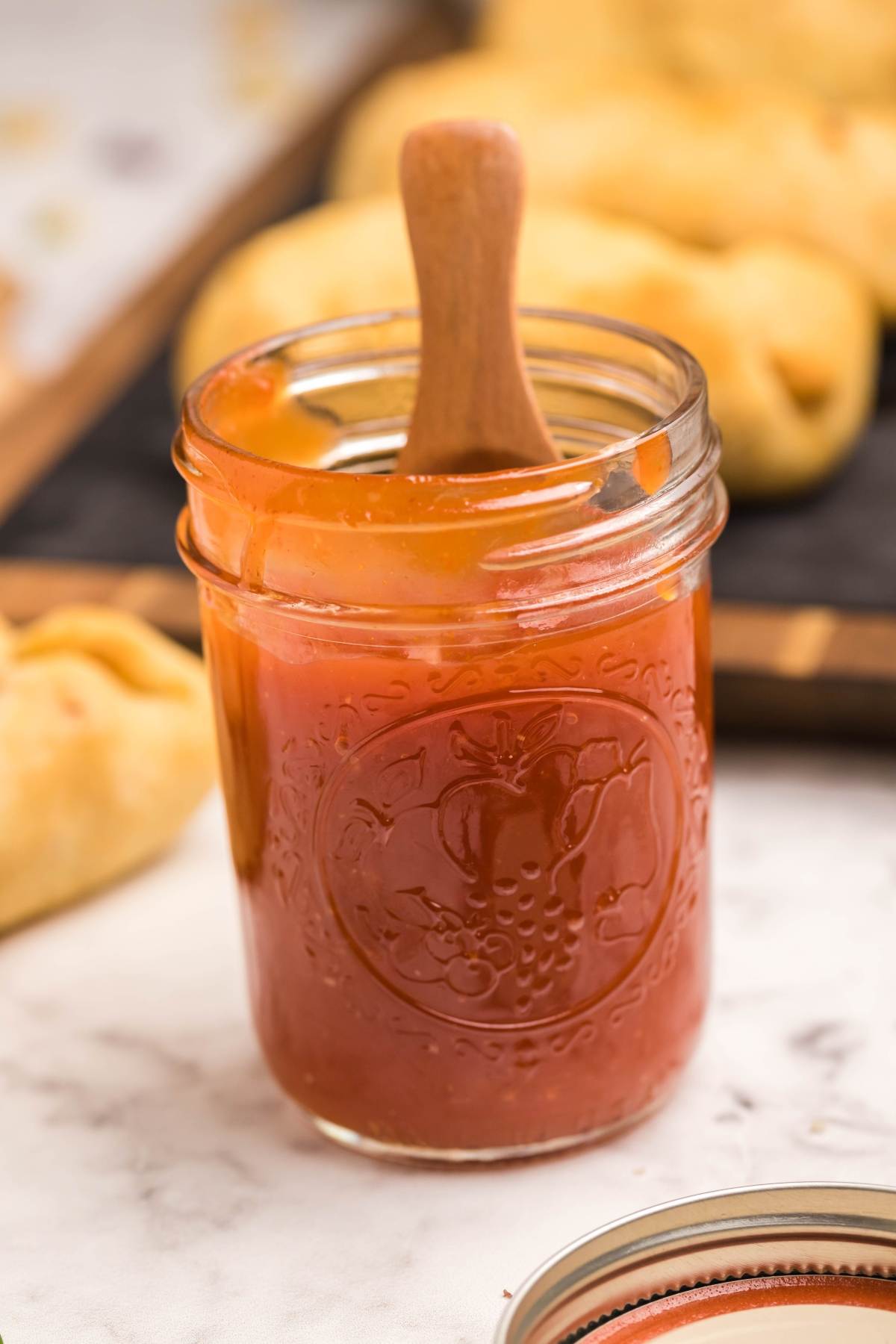 A glass jar of peach jam, perfect as a sugar free sweet and sour sauce, with a wooden spoon inside, sits on a marble surface.