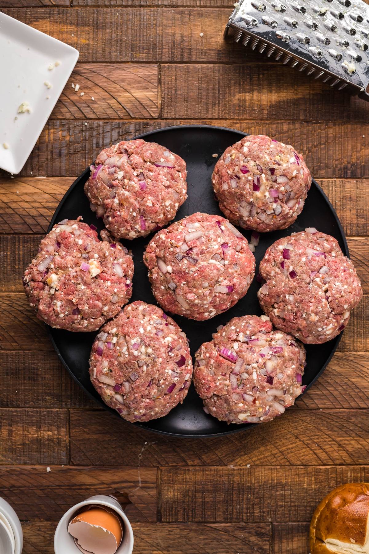 A plate of six raw bison burgers with chopped onions sits on a wooden surface, surrounded by cooking utensils—perfect for grilling or serving on a pretzel bun.