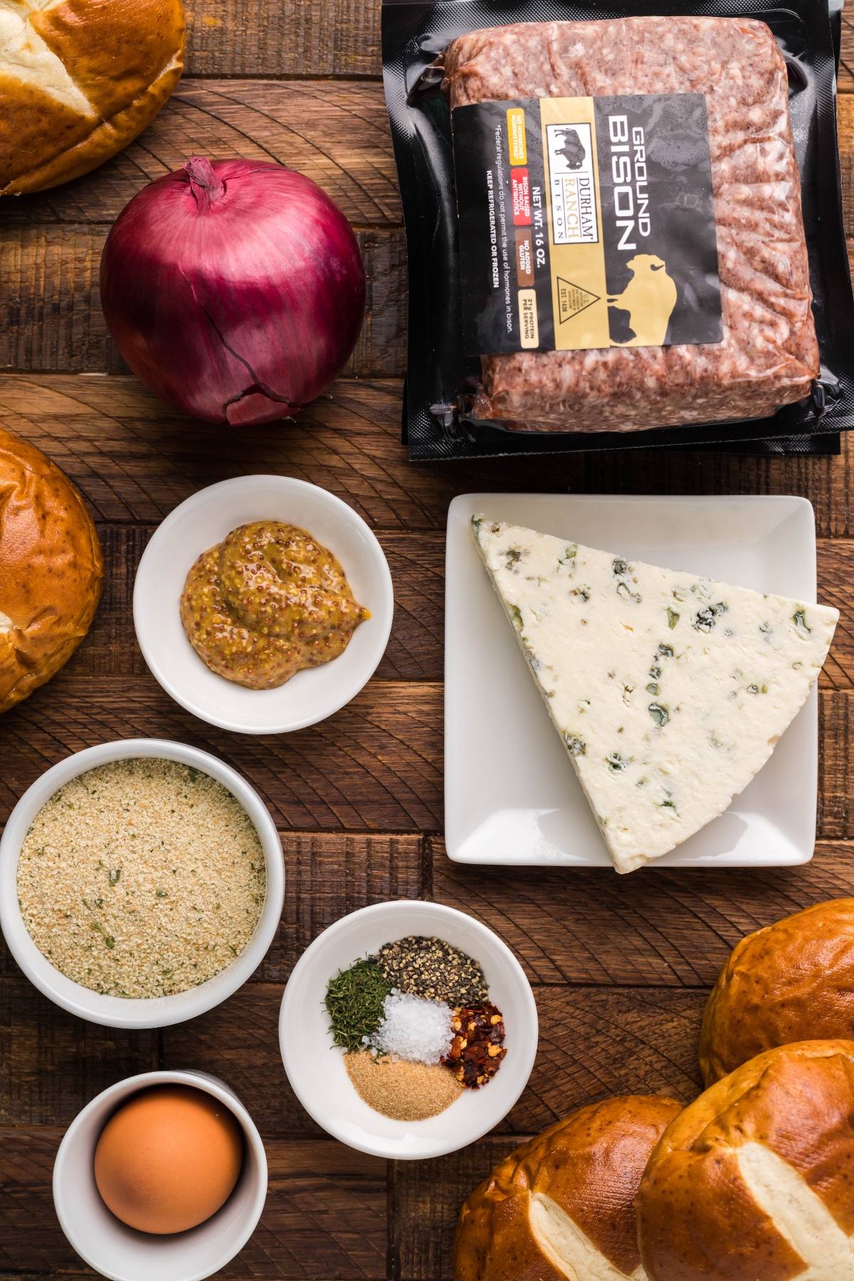 Ground bison, blue cheese, onion, buns, egg, breadcrumbs, spices, and mustard come together for savory Gorgonzola Buffalo Burgers—perfectly paired with a soft pretzel bun on a rustic wooden surface.