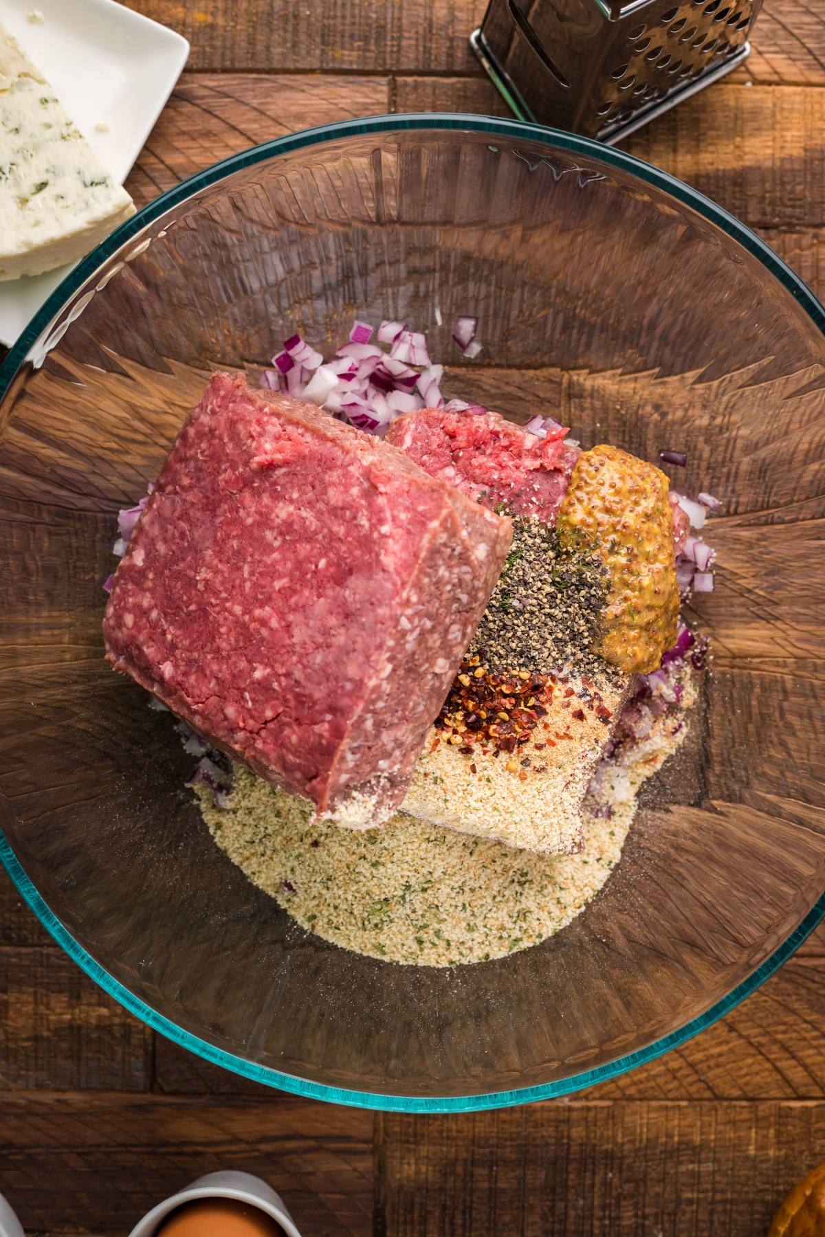 Ground beef, spices, chopped onions, breadcrumbs, and crumbled blue cheese in a glass bowl on a wooden surface—ready to mix the perfect Gorgonzola Buffalo Burgers.