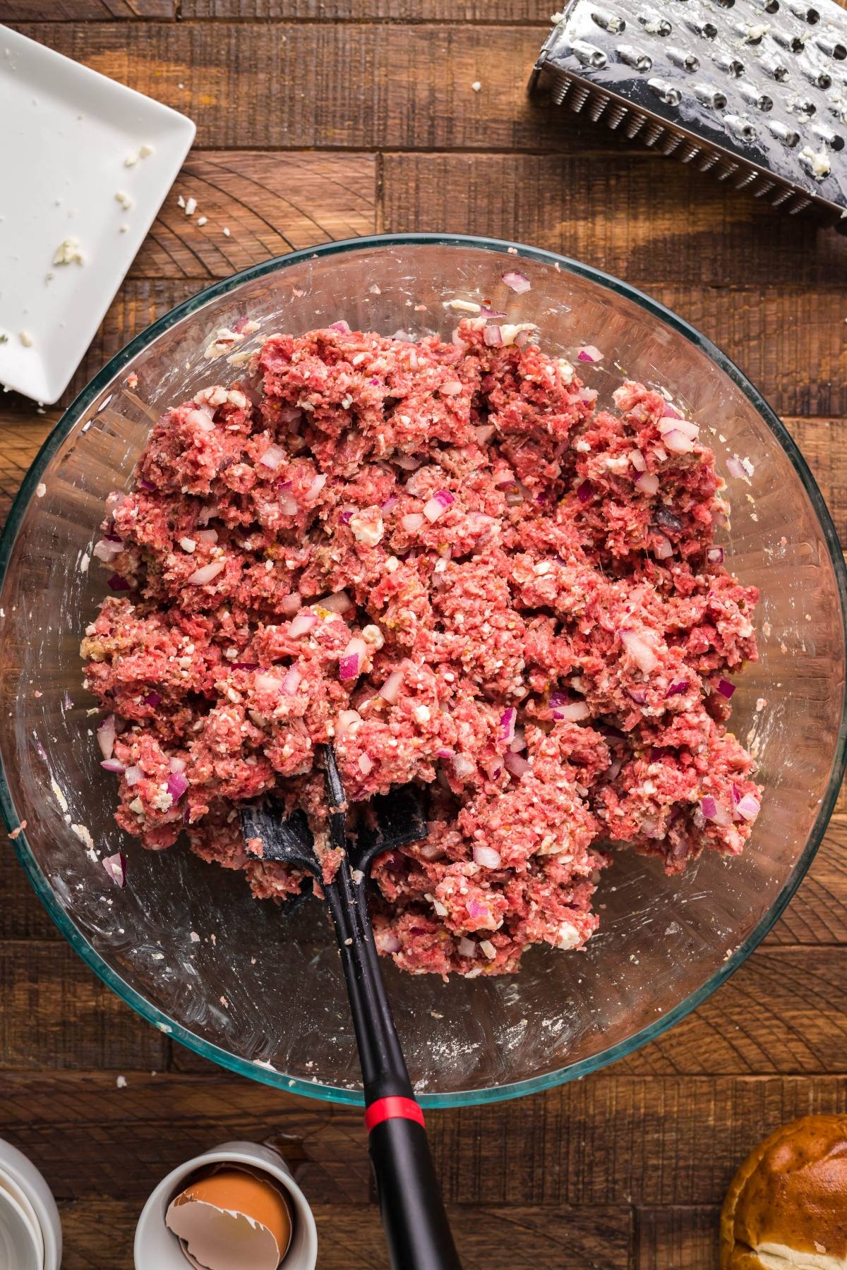 Ground meat mixed with chopped onions and seasonings in a glass bowl with a black spoon on a wooden surface, perfect for making flavorful Gorgonzola Buffalo Burgers.