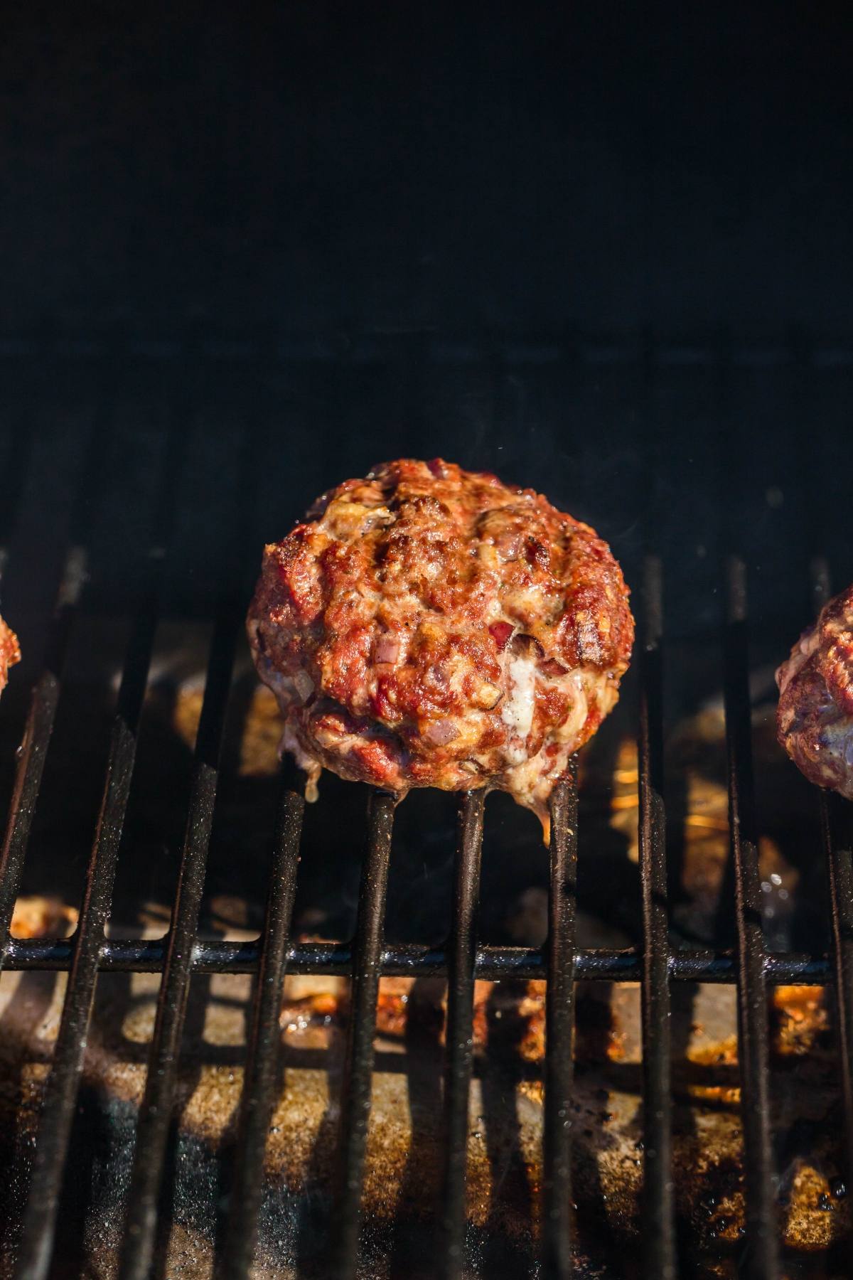 A beef burger patty grilling on a barbecue, with grill marks and a smoky background, ready to be topped with blue cheese or gorgonzola and served on a warm pretzel bun.