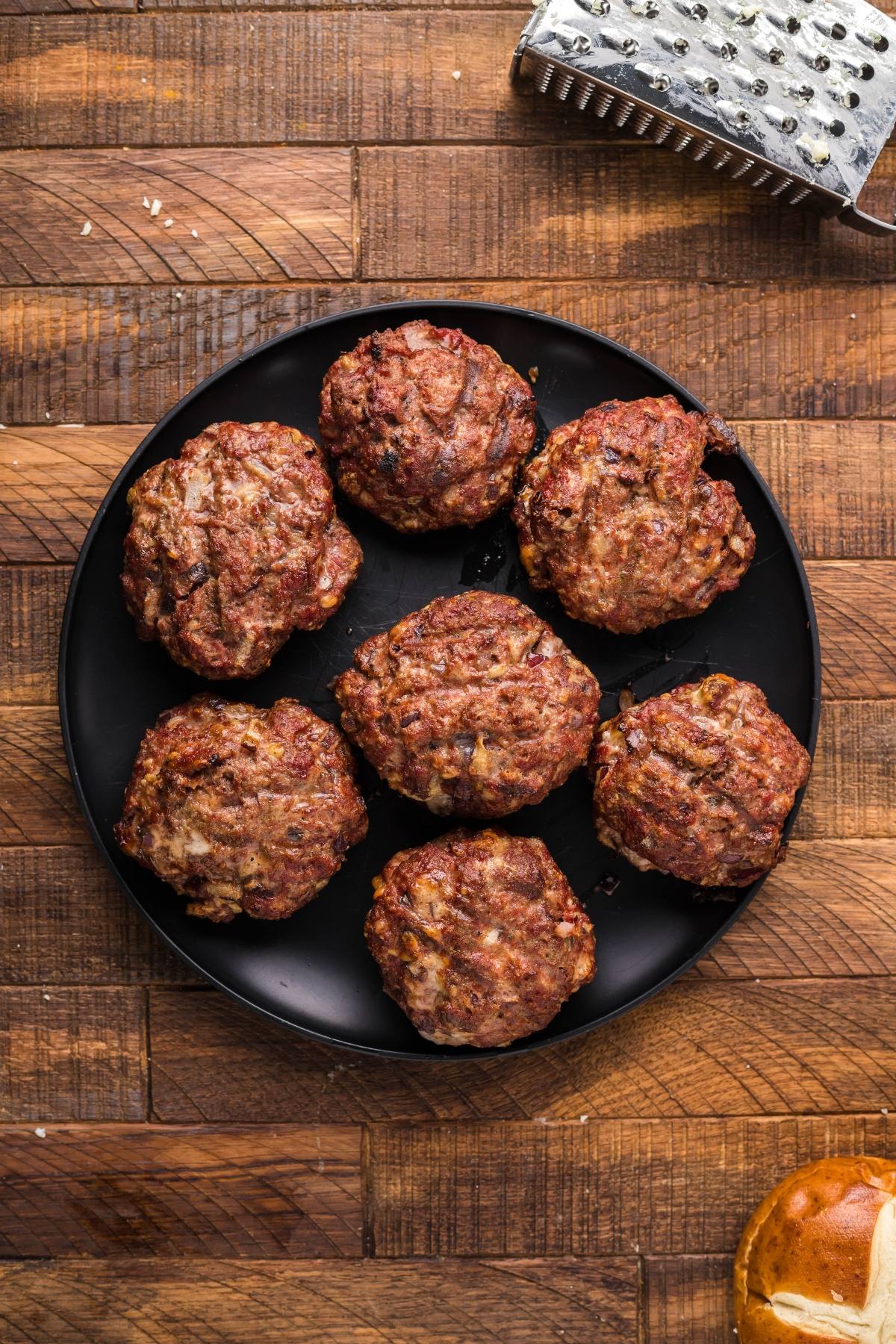 Seven cooked Grilled buffalo burger patties on a black plate, set on a wooden table with a grater and a burger bun nearby—perfect for making Gorgonzola Buffalo Burgers.
