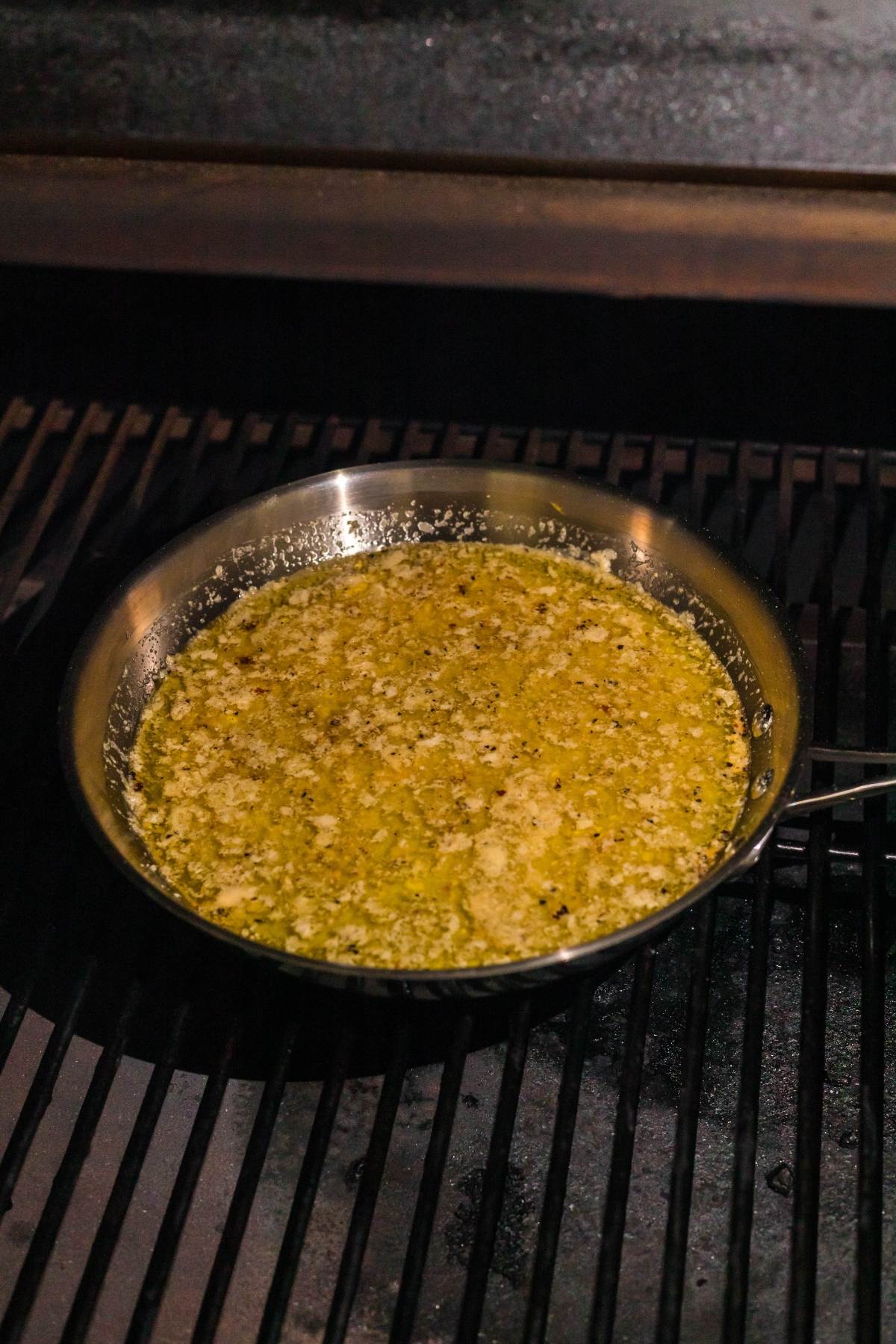A metal pan with melted butter, herbs, and spices sits on a grill, ready to baste delicious BBQ scallop skewers as they cook.