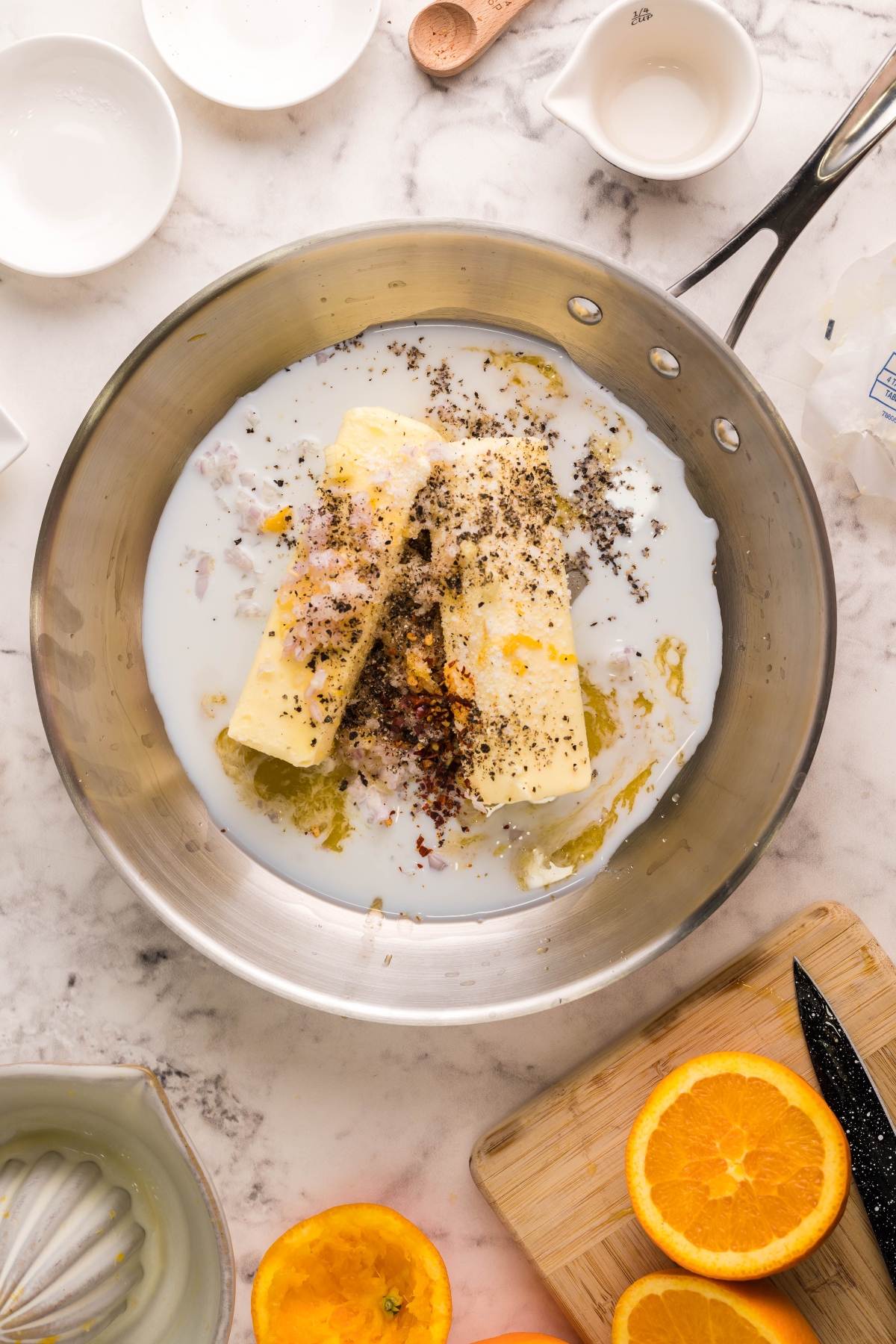 Butter, spices, and milk simmer in a saucepan on a marble countertop with sliced oranges and kitchen tools nearby—perfect for drizzling over BBQ scallop skewers or pairing with a Citrus Garlic Smoked Butter Sauce.