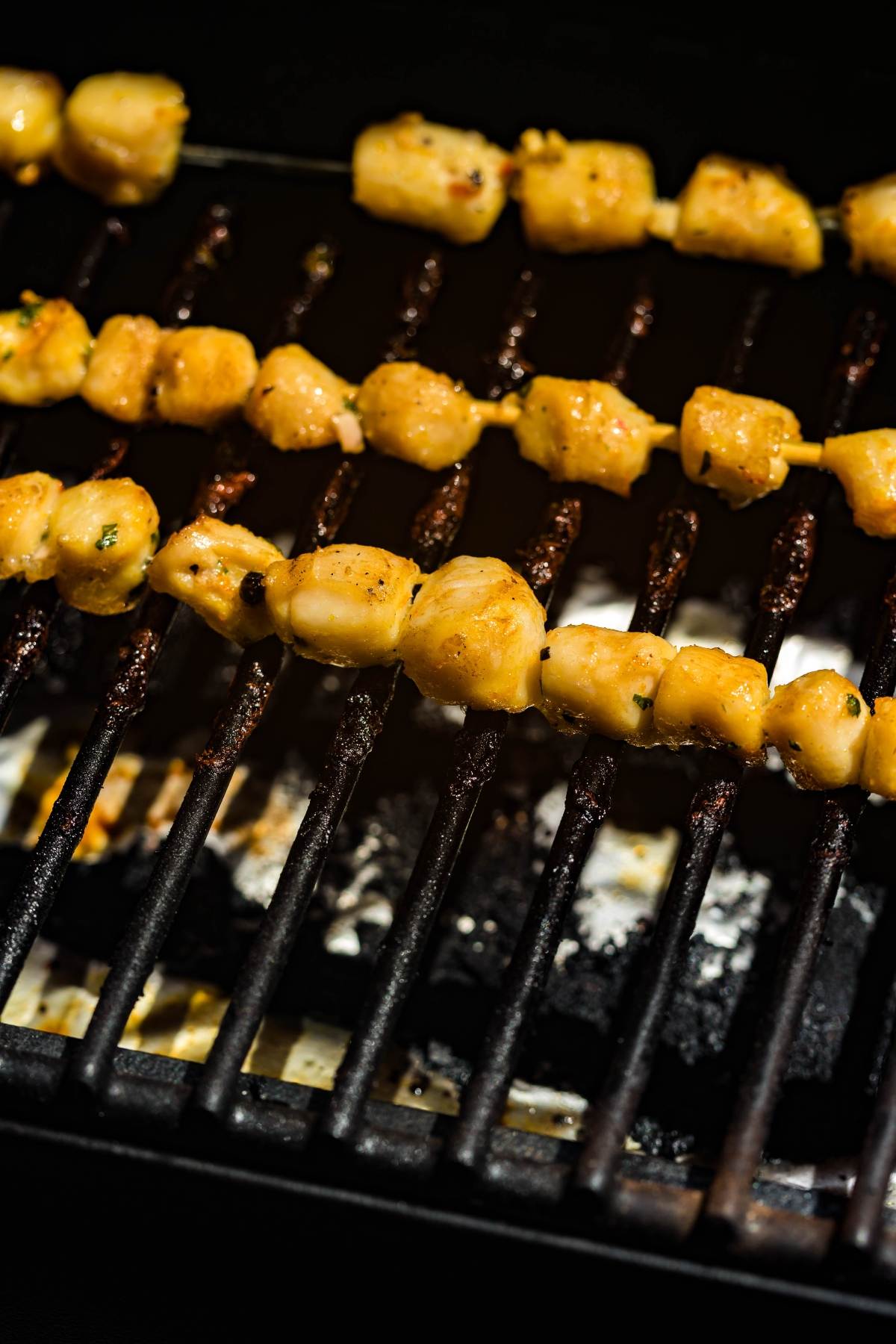 Chicken skewers cooking on a grill, with golden-brown pieces over hot metal grates, share the space with BBQ scallop skewers for a delicious mixed grill feast.
