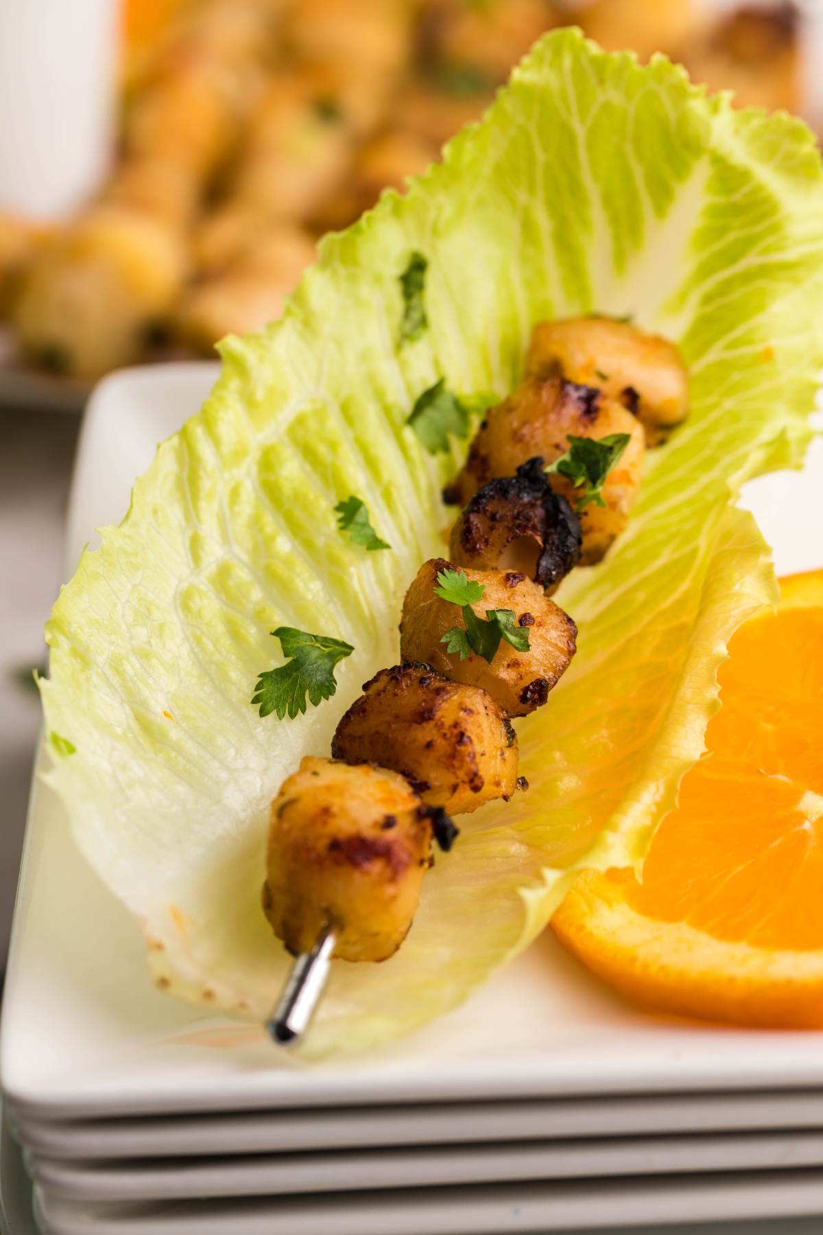 Grilled skewered chicken on a lettuce leaf with cilantro and an orange slice, served beside BBQ scallop skewers and finished with a drizzle of Citrus Garlic Smoked Butter Sauce on a white plate.