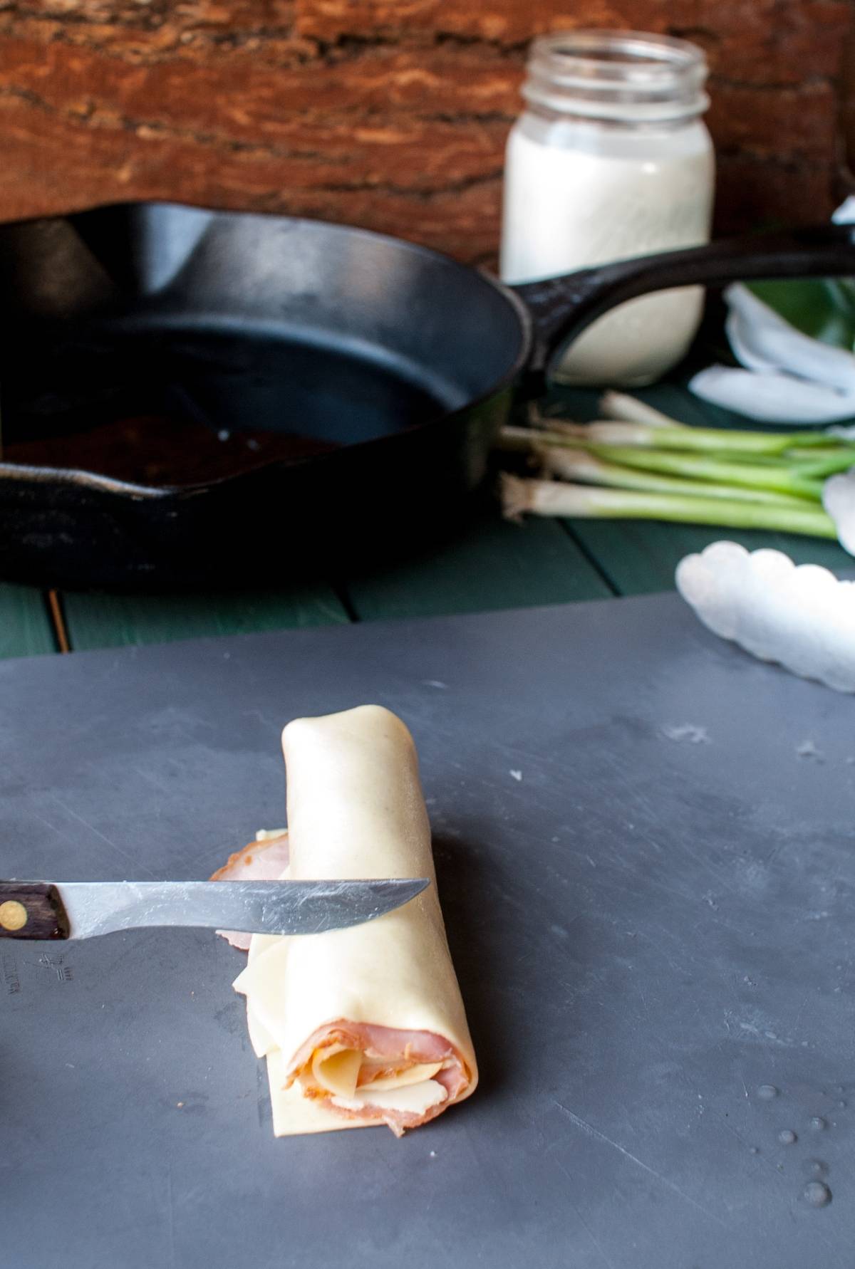 A knife beside a rolled ham and cheese wrap on a cutting board, resembling pasta pinwheels, with a skillet and jar in the background.