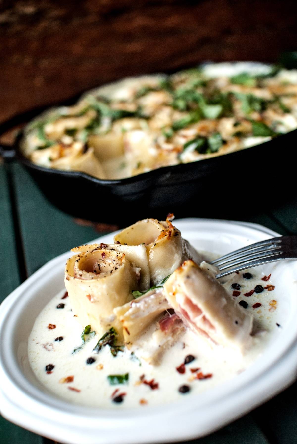 Rolled pasta pinwheels with creamy white sauce and herbs on a white plate; casserole dish in background, reminiscent of Ham and Provolone Rotolo – Lasagna Roll Ups.