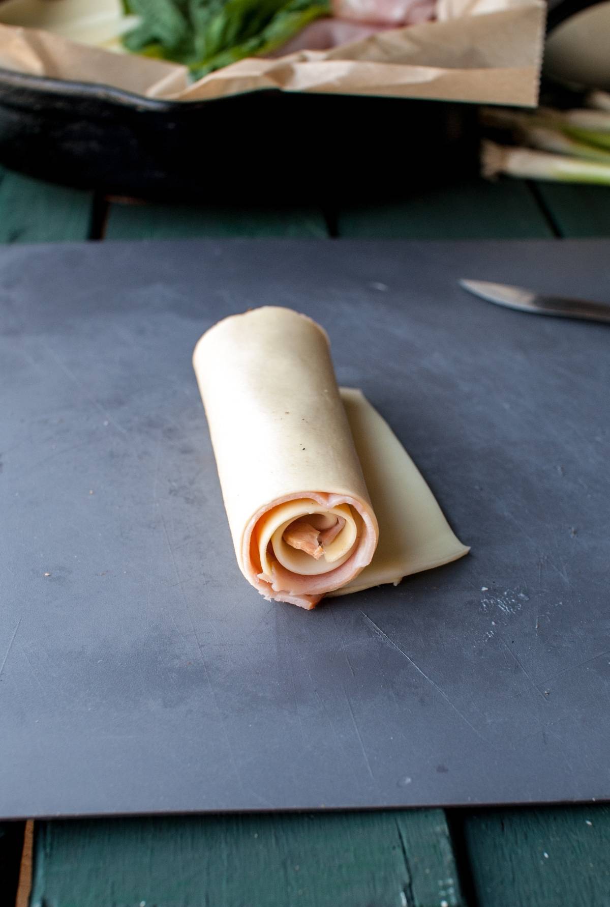 A slice of cheese and deli meat rolled up on a gray cutting board, resembling Ham and Provolone Rotolo – Lasagna Roll Ups, with a knife in the background.