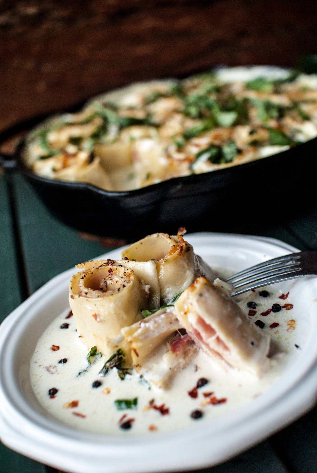 Pasta rolls with ham and creamy white sauce on a plate, featuring Ham and Provolone Rotolo – Lasagna Roll Ups, with a baked dish of pasta roses in the background.