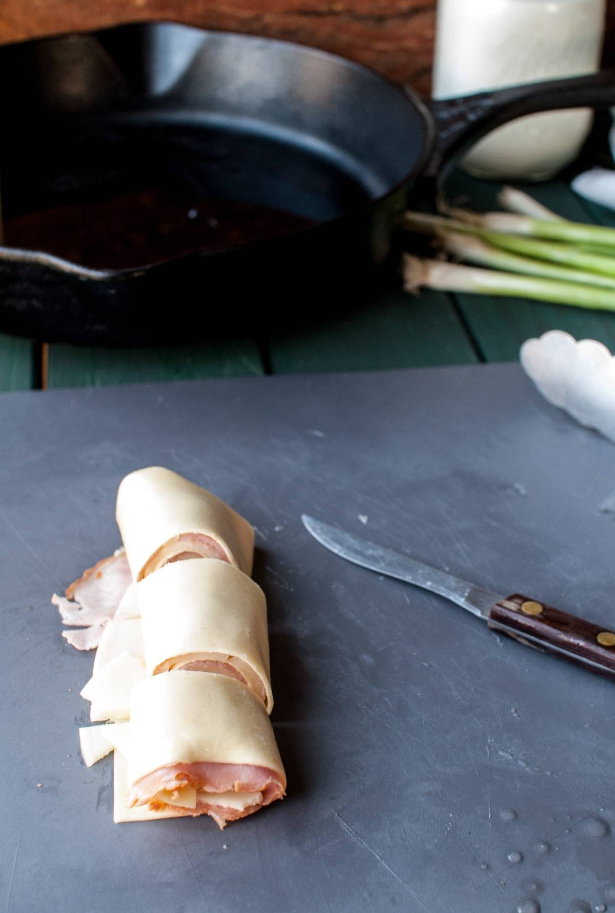 Four unbaked ham and cheese crescent rolls resembling pasta pinwheels rest on a gray surface beside a knife and a black skillet.