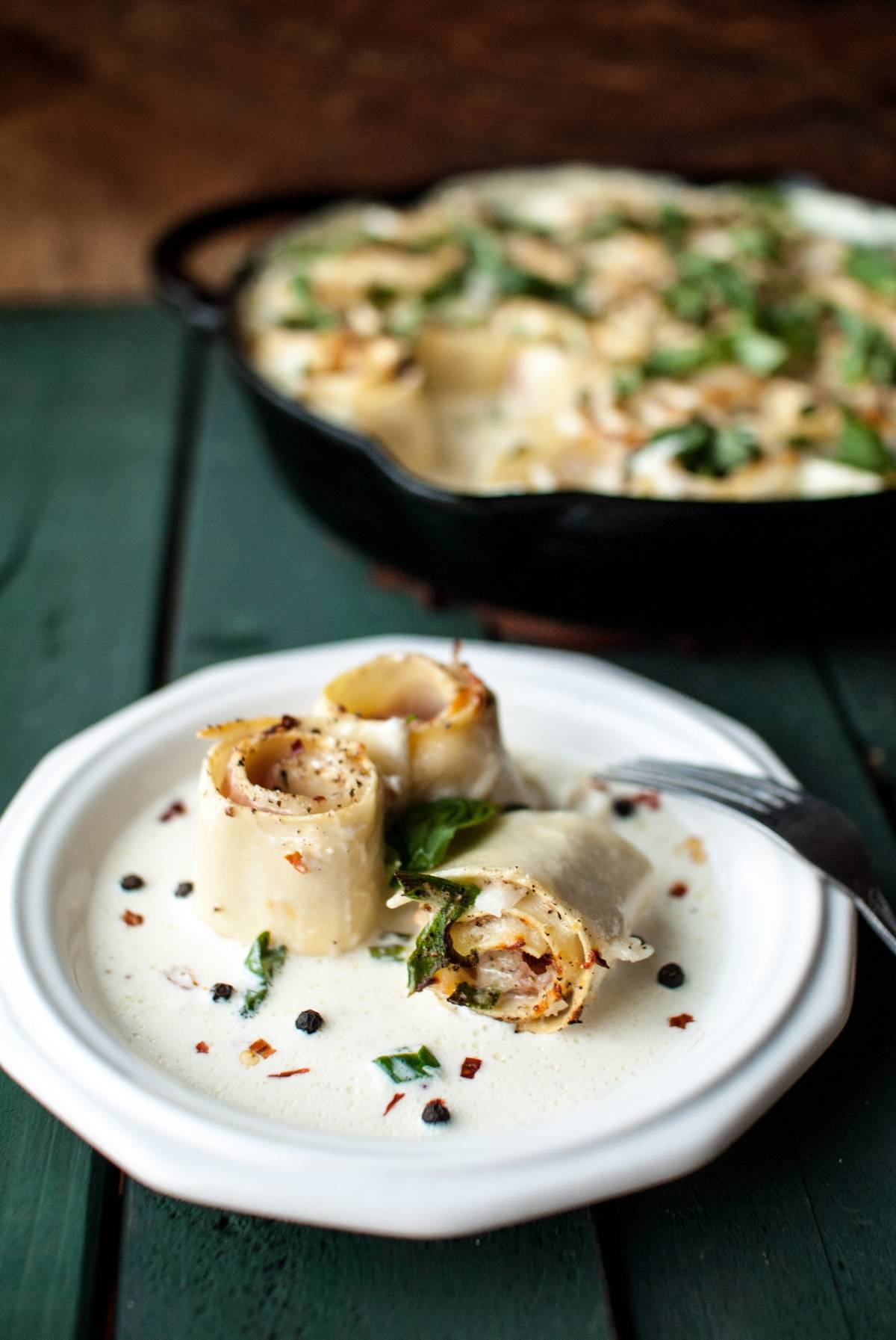 Two Lasagna Roll Ups with creamy sauce and herbs rest on a white plate, with more Ham and Provolone Rotolo – Lasagna Roll Ups visible in a skillet in the background.
